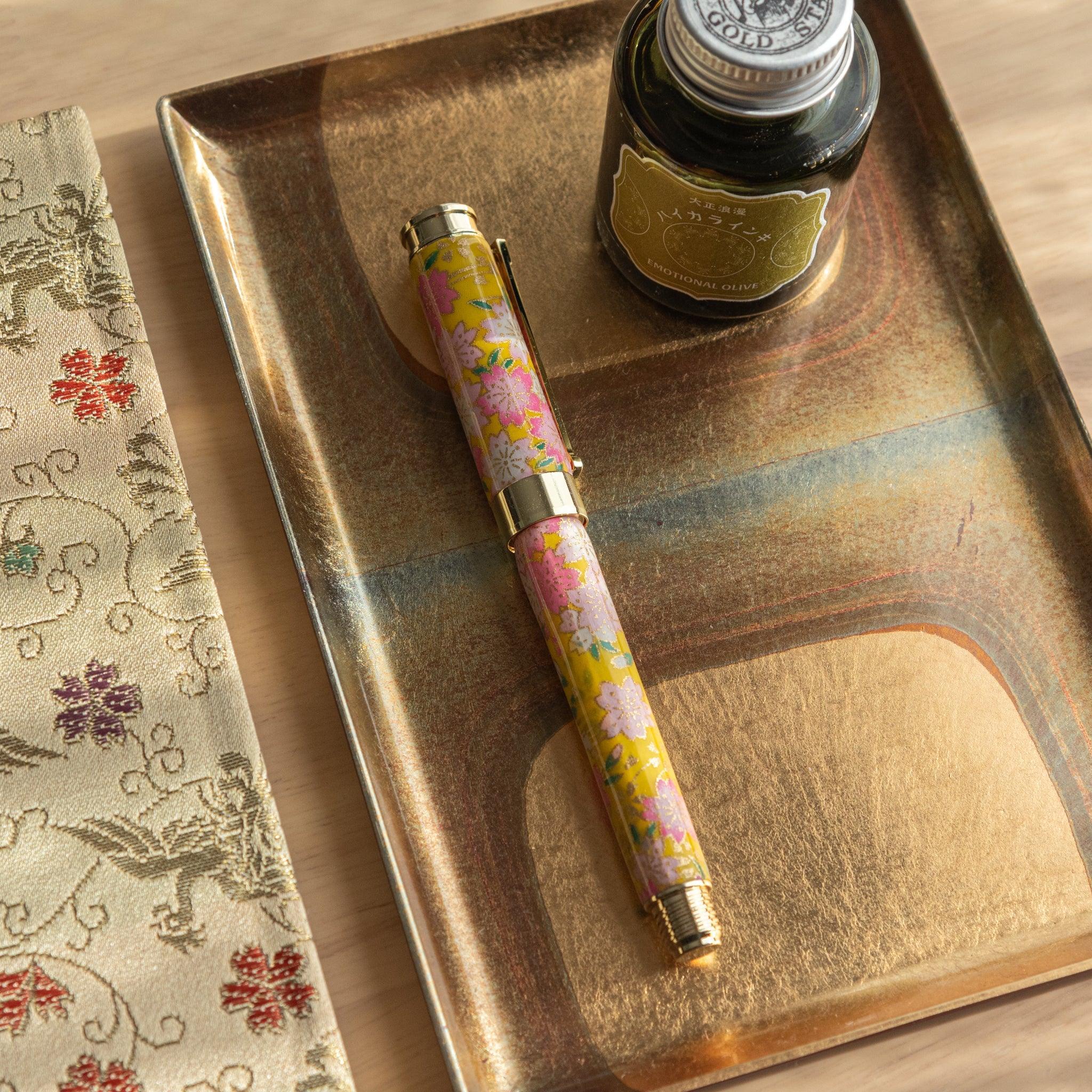 Closed Japanese fountain pen with sakura and canola pattern in pink and yellow, displayed with ink on a golden tray