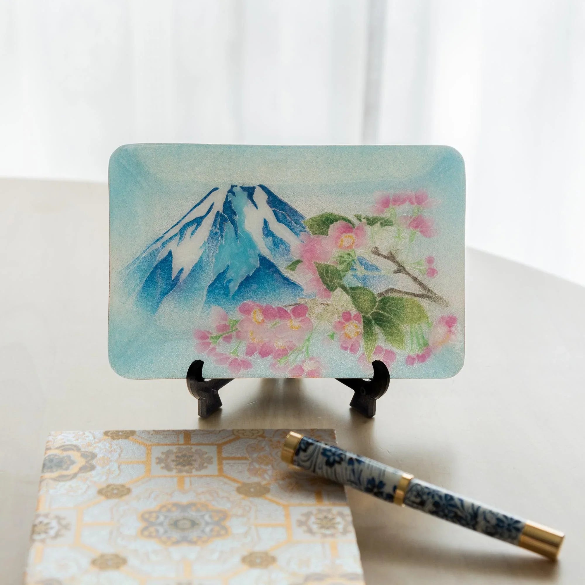Japanese shippo ware pen tray on display stand showing Mt. Fuji and pink sakura design, blue floral pen and notebook below