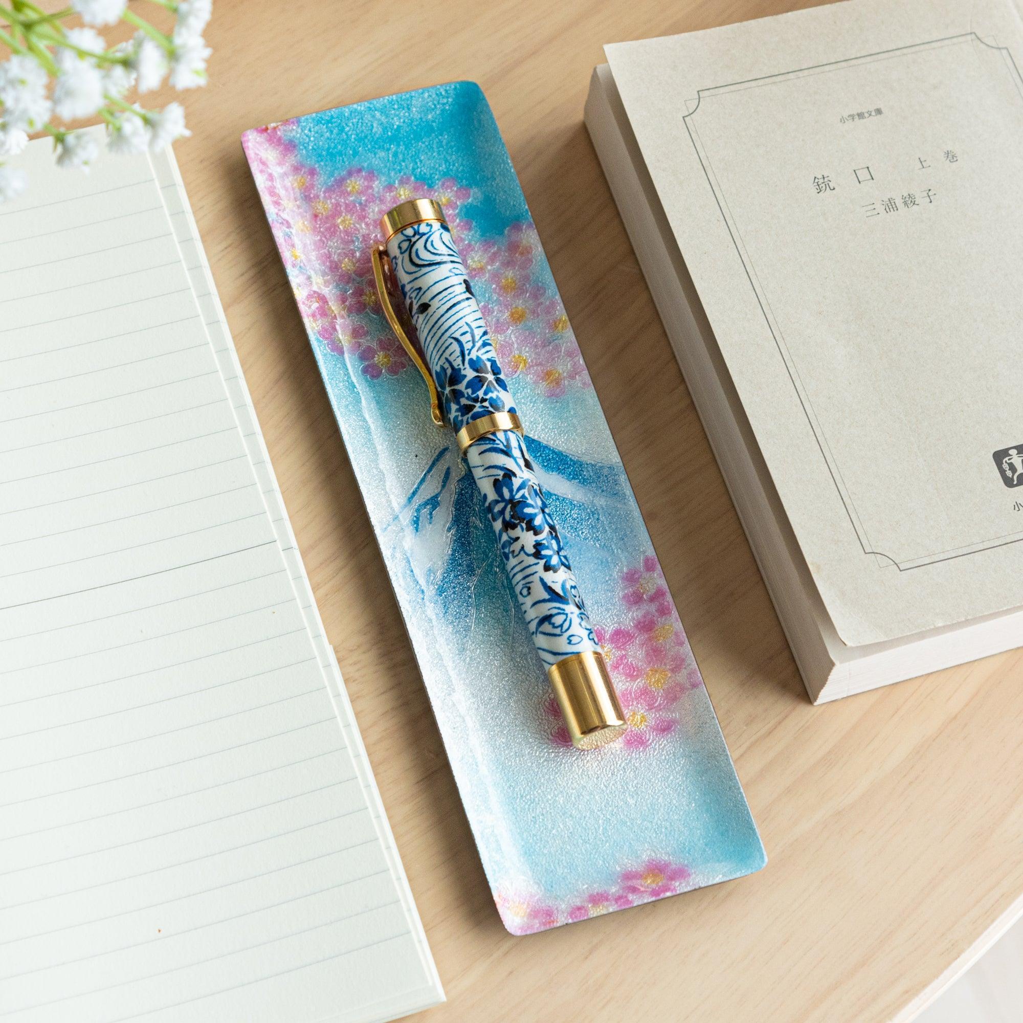 Decorative tray featuring Mount Fuji and cherry blossoms, holding a white and blue floral pen with gold accents, placed on a wooden desk.