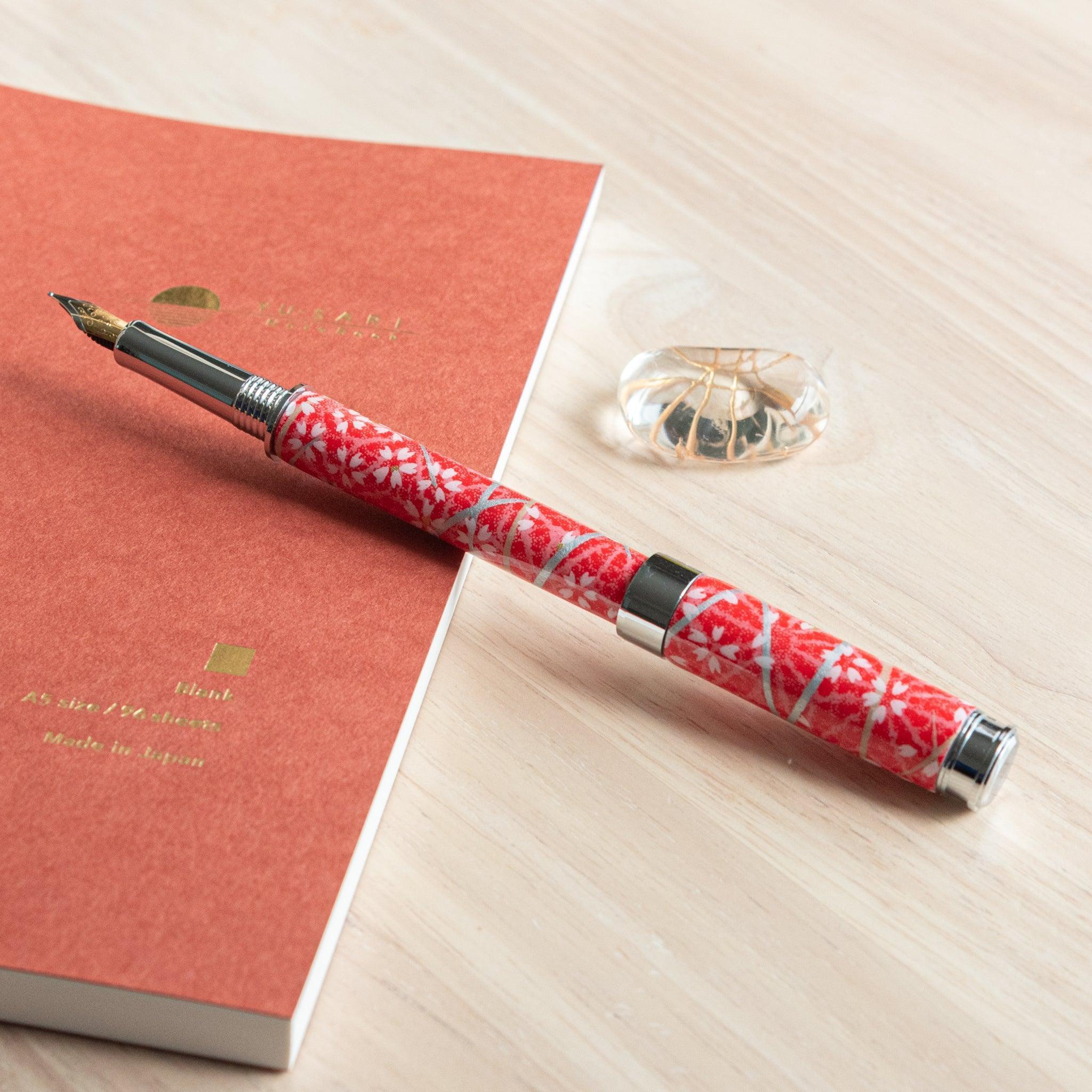 A Japanese stationery red-patterned fountain pen with its cap removed, revealing the nib, lying next to an orange notebook on a wooden surface.