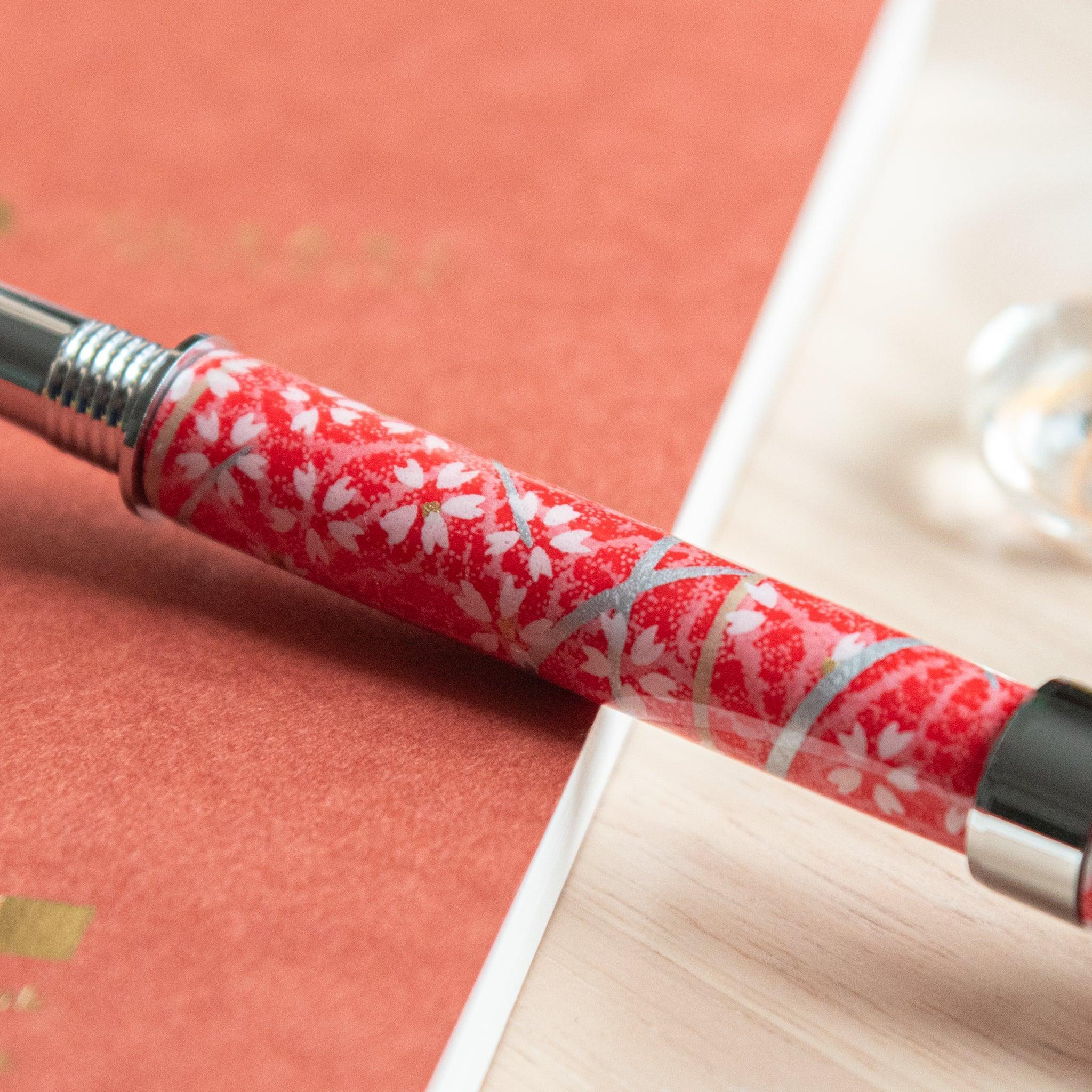 A Japanese stationery fountain pen with red floral patterns and silver details, resting on an orange notebook.