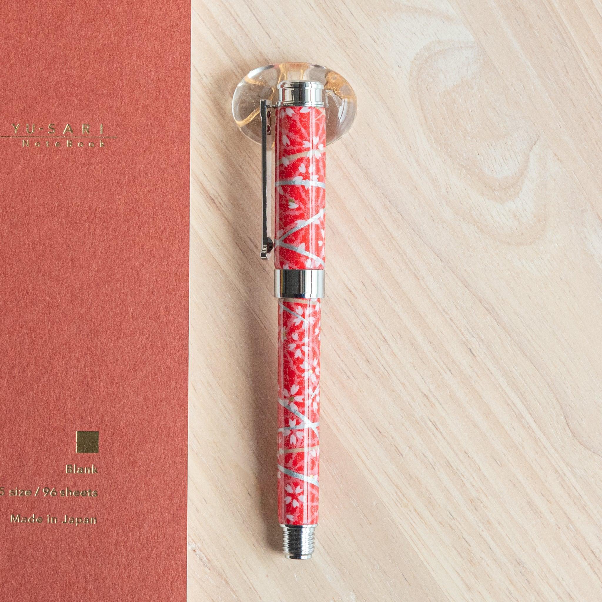 A Japanese stationery red-patterned fountain pen with silver accents placed vertically on a wooden surface beside an orange notebook.