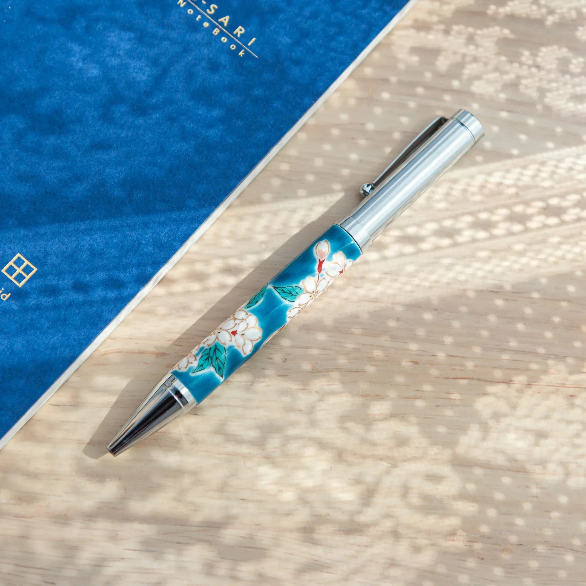 Japanese Ballpoint Pen with Kutani cherry blossom design, silver clip visible on left beside blue notebook.