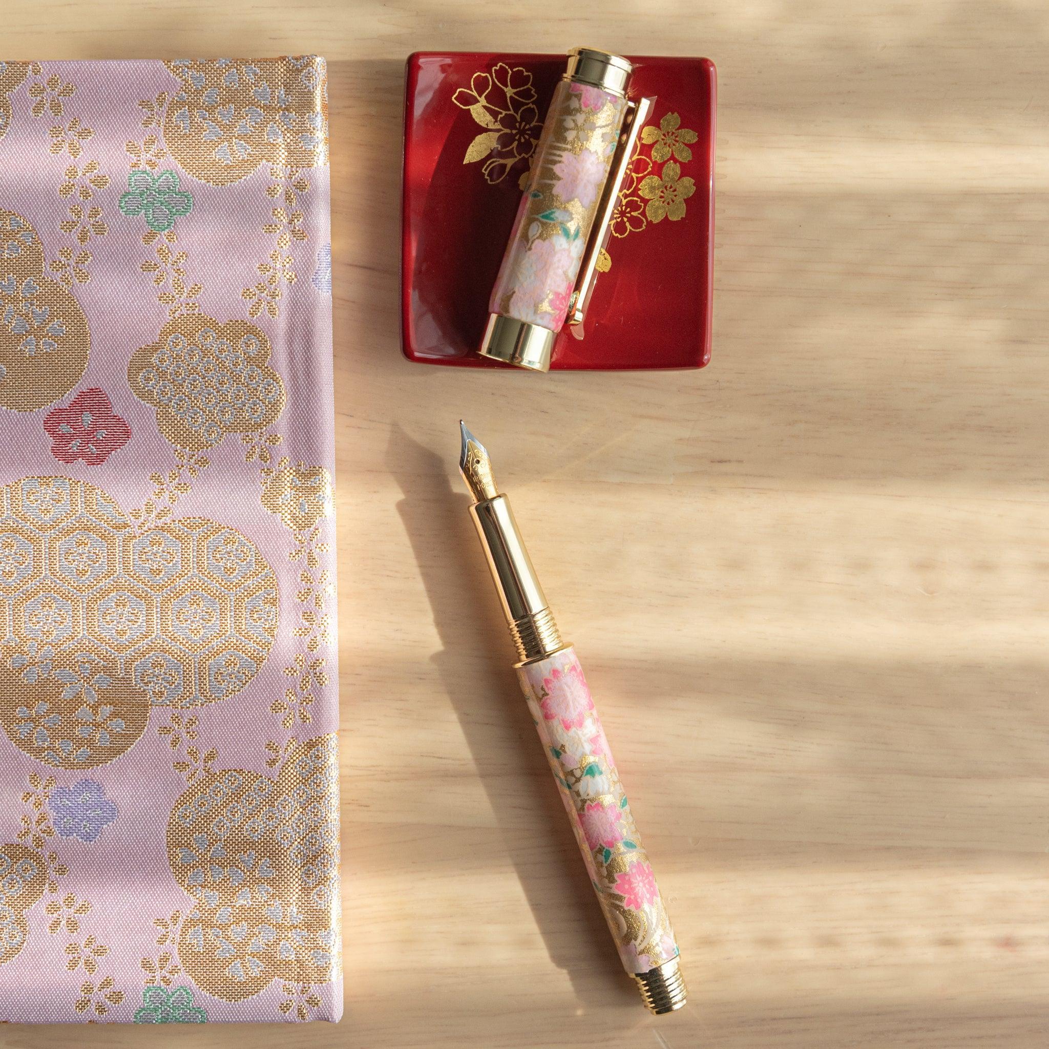 Open pink sakura-patterned fountain pen with gold details, displayed with cap on a red tray and golden-patterned fabric.