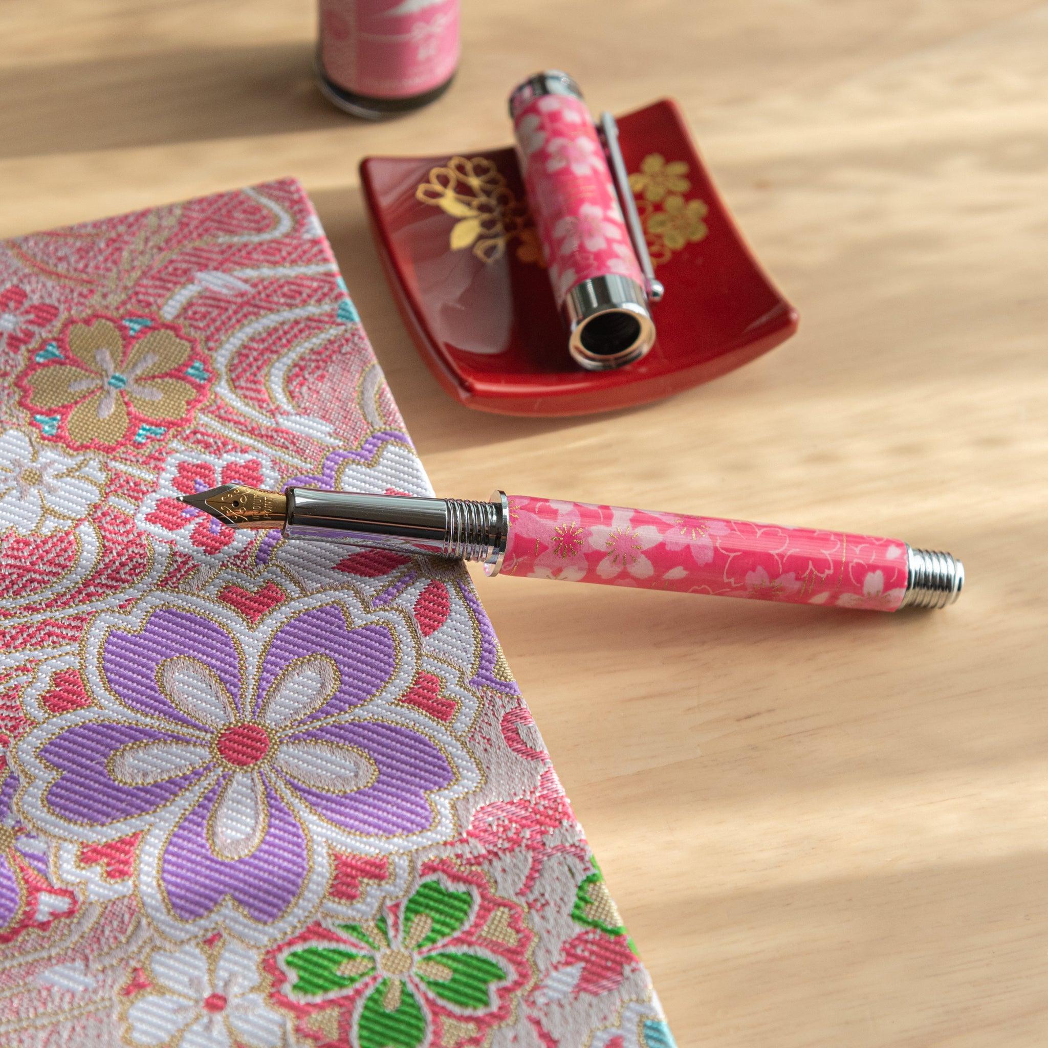 A pink floral Japanese stationery fountain pen disassembled on a table with a colorful fabric and red dish in the background.