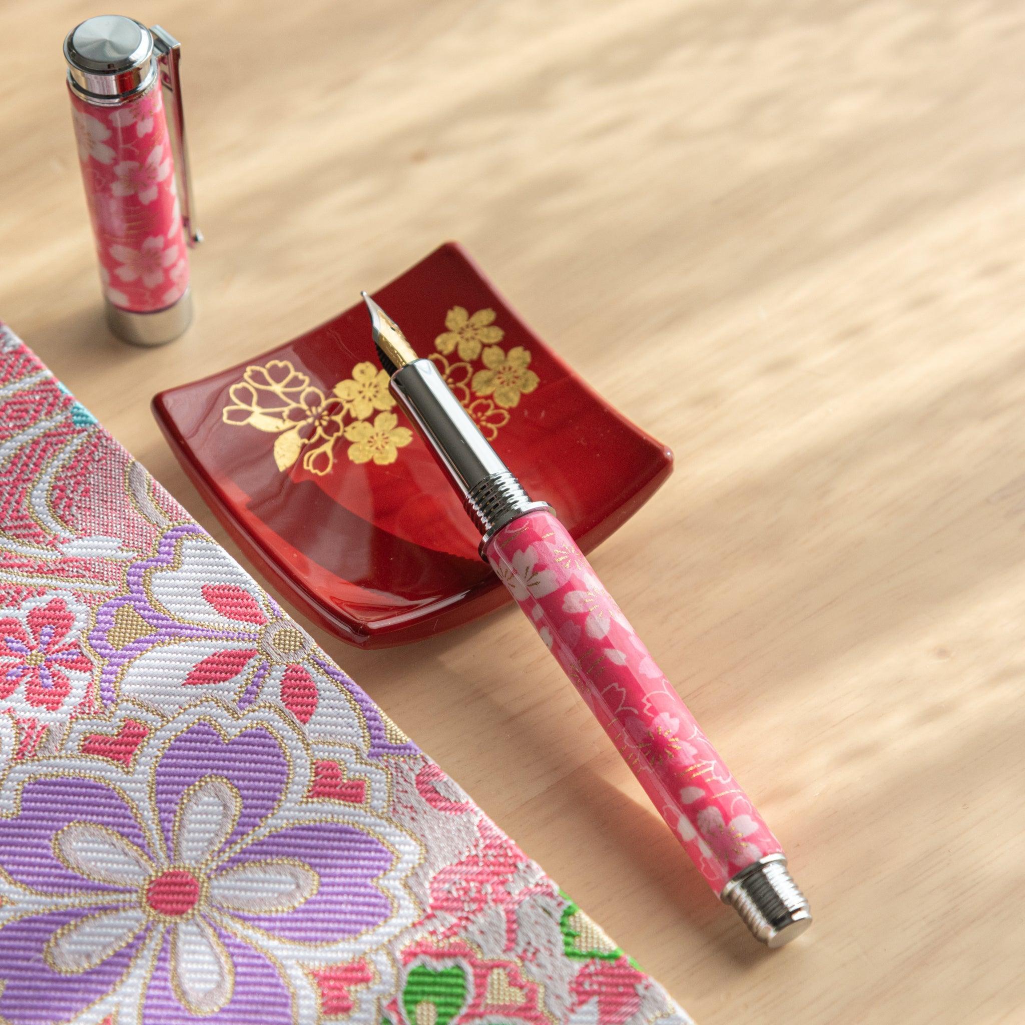 A pink floral Japanese stationery fountain pen with its nib exposed, placed near a red dish and colorful fabric.