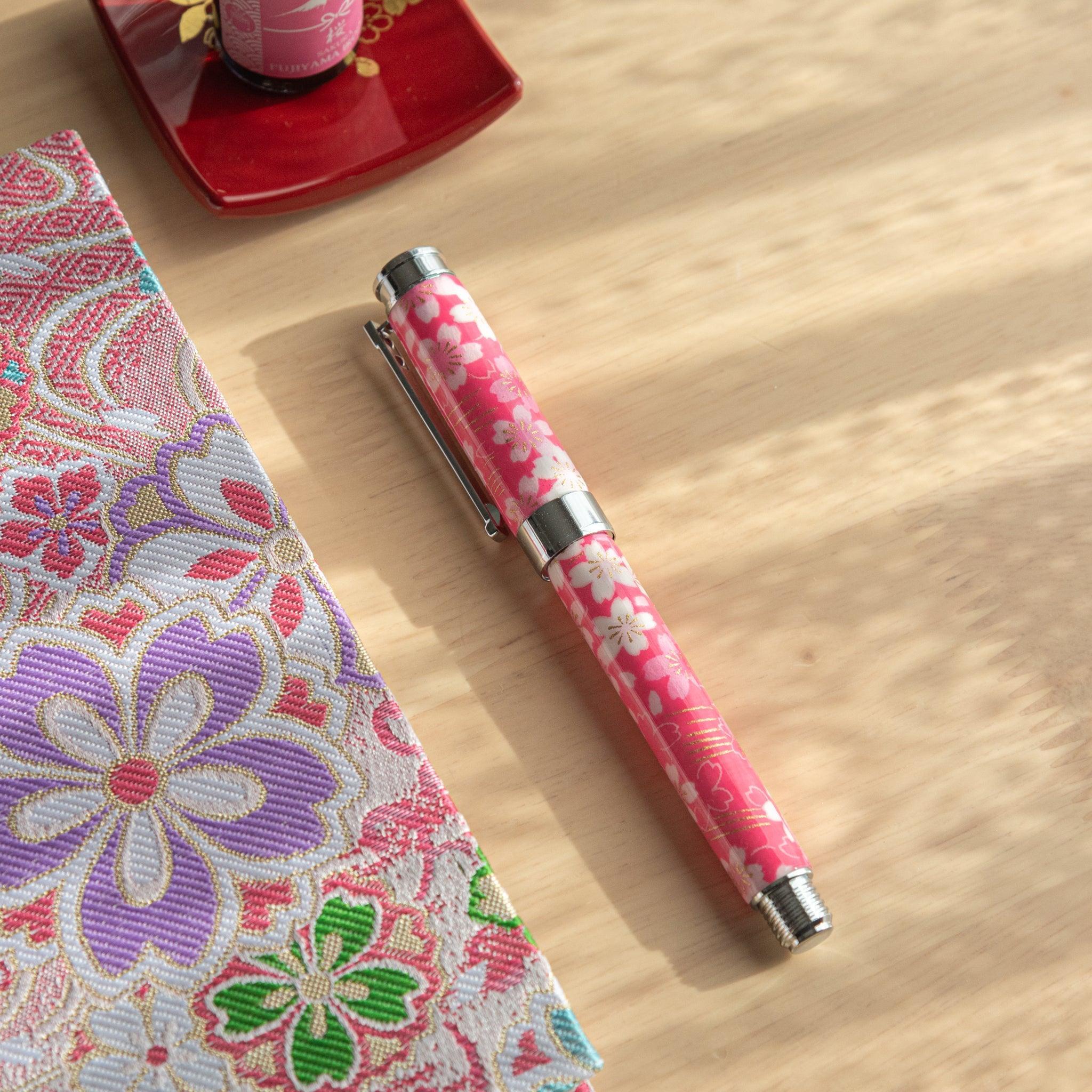 A pink floral Japanese stationery fountain pen placed on a table next to a colorful fabric and a red dish under sunlight.