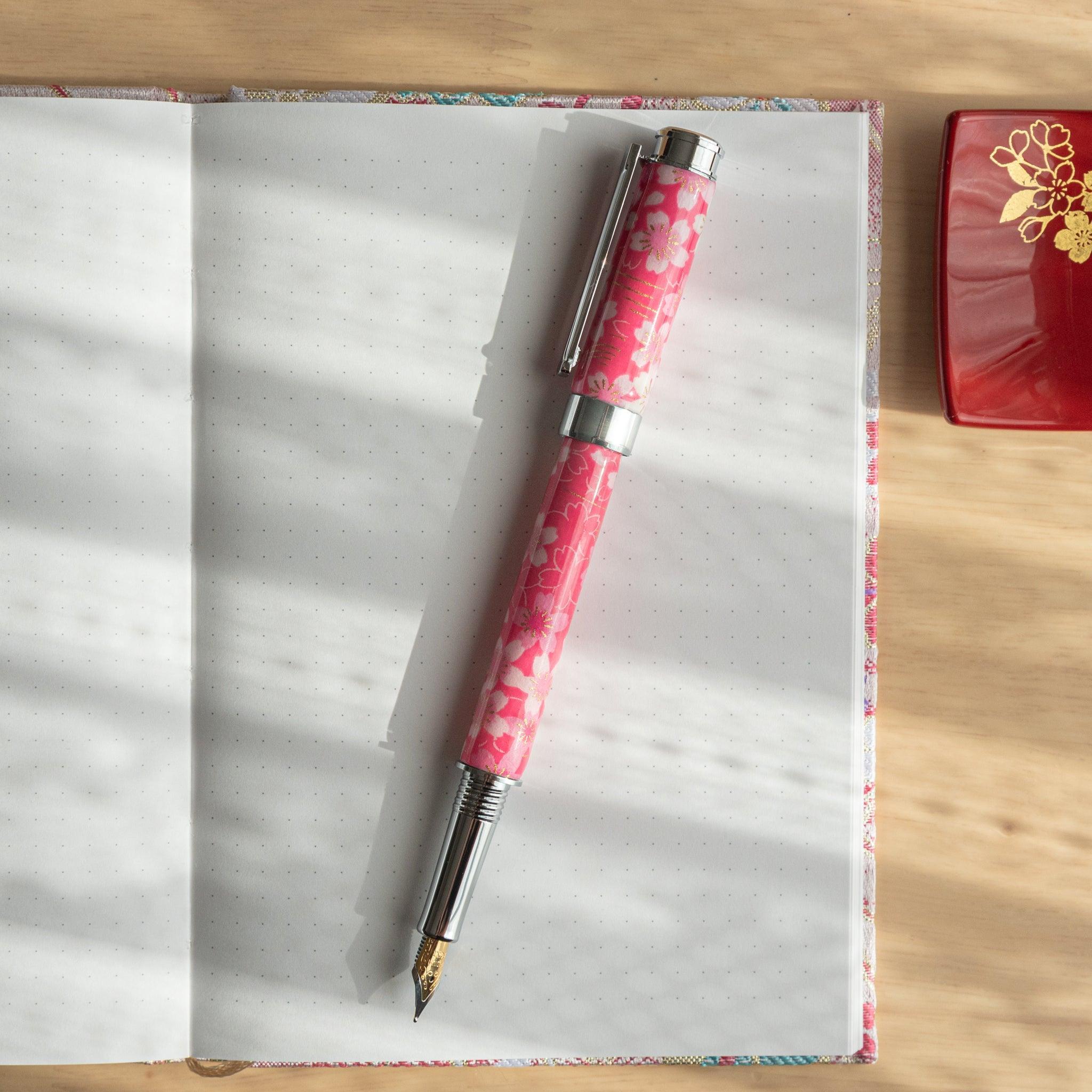 A pink floral Japanese stationery fountain pen placed on an open notebook under soft sunlight.