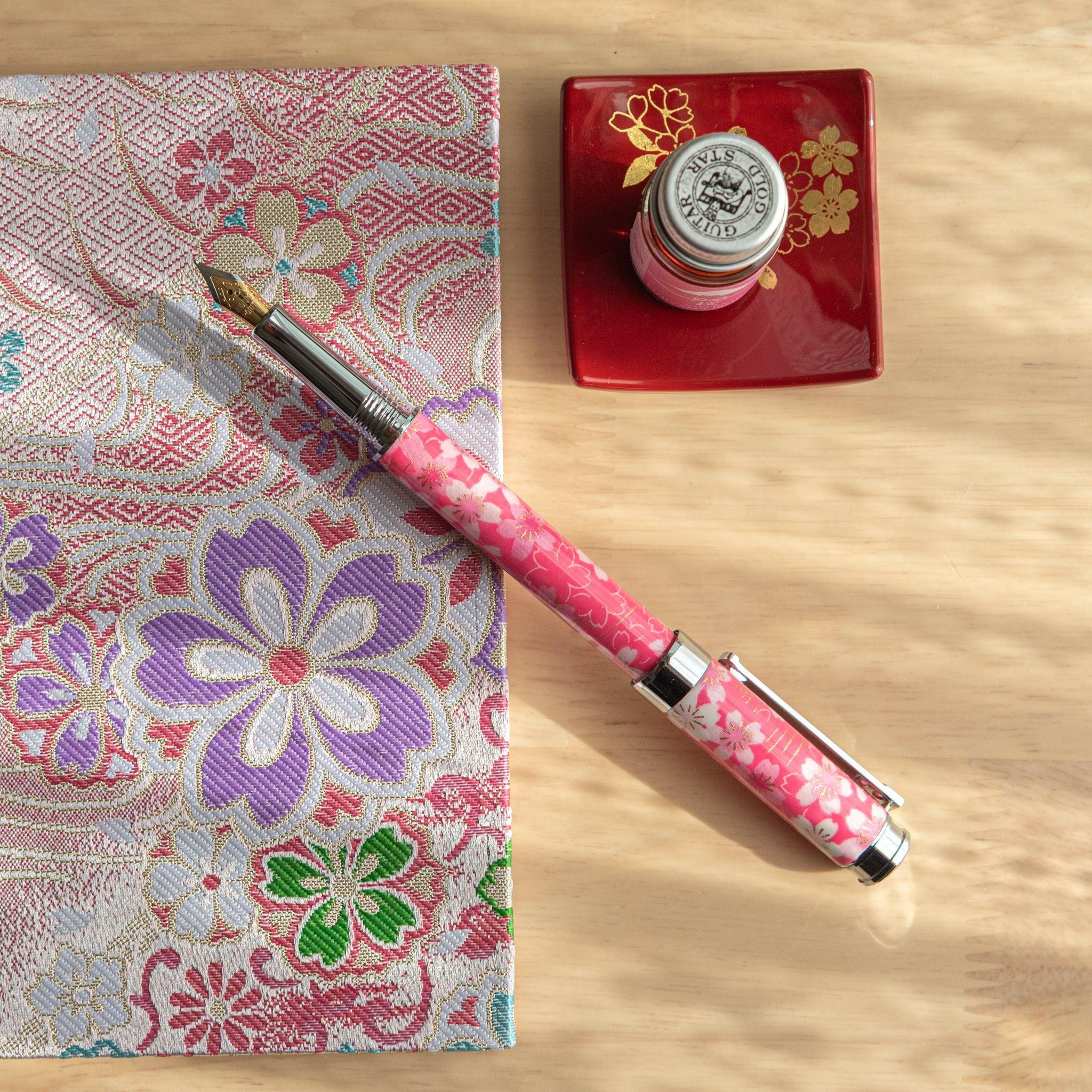 A pink floral Japanese stationery fountain pen resting on colorful fabric with a red dish and ink bottle nearby.