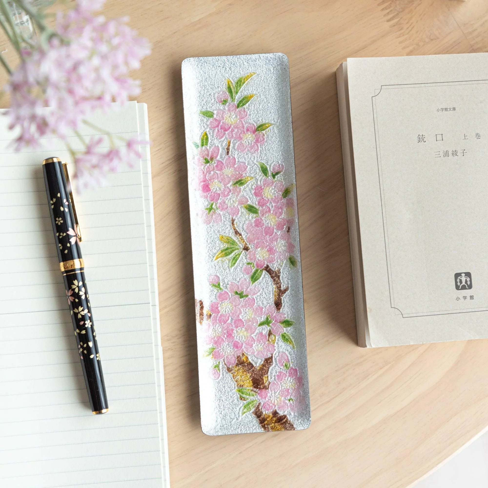 Silver enamel slim pen tray with pink sakura and green leaf design, on wood desk with black sakura fountain pen and notebook