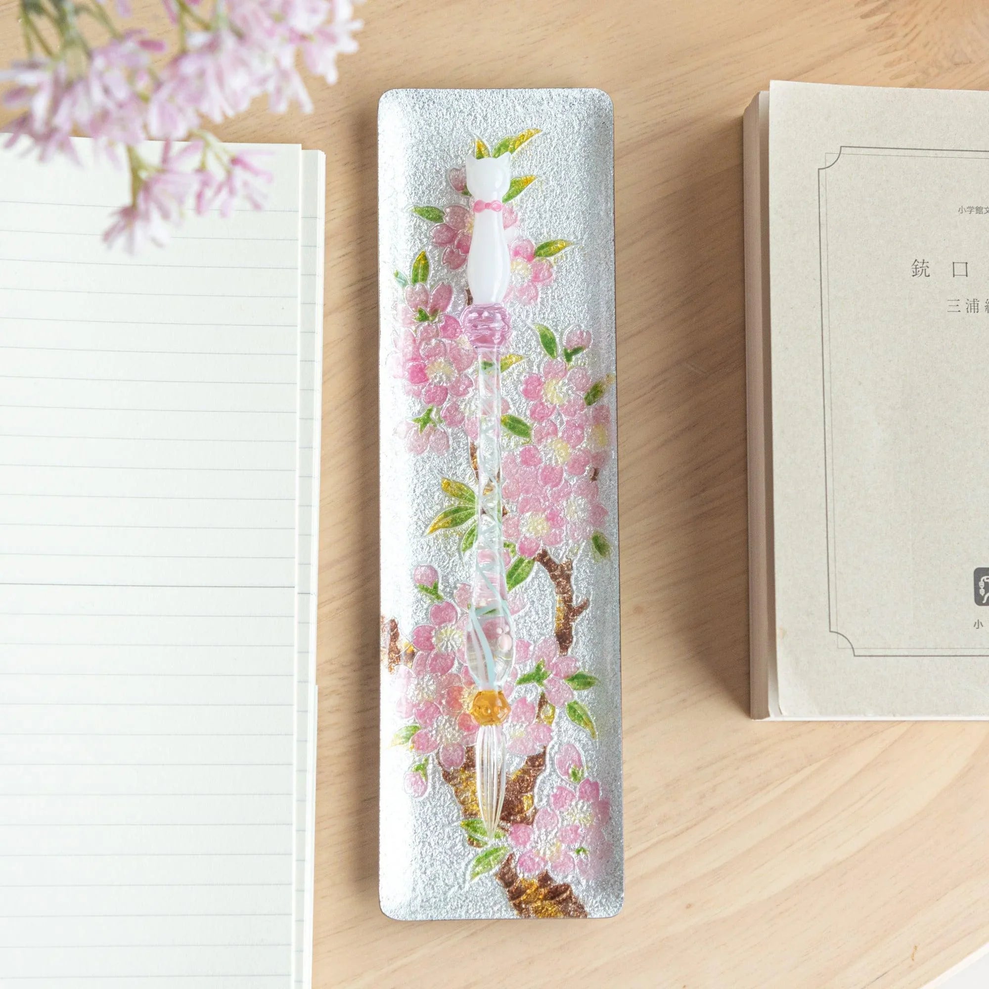 Silver enamel slim pen tray with pink sakura design, overhead view, glass pen with pink resting on tray, notebook and book
