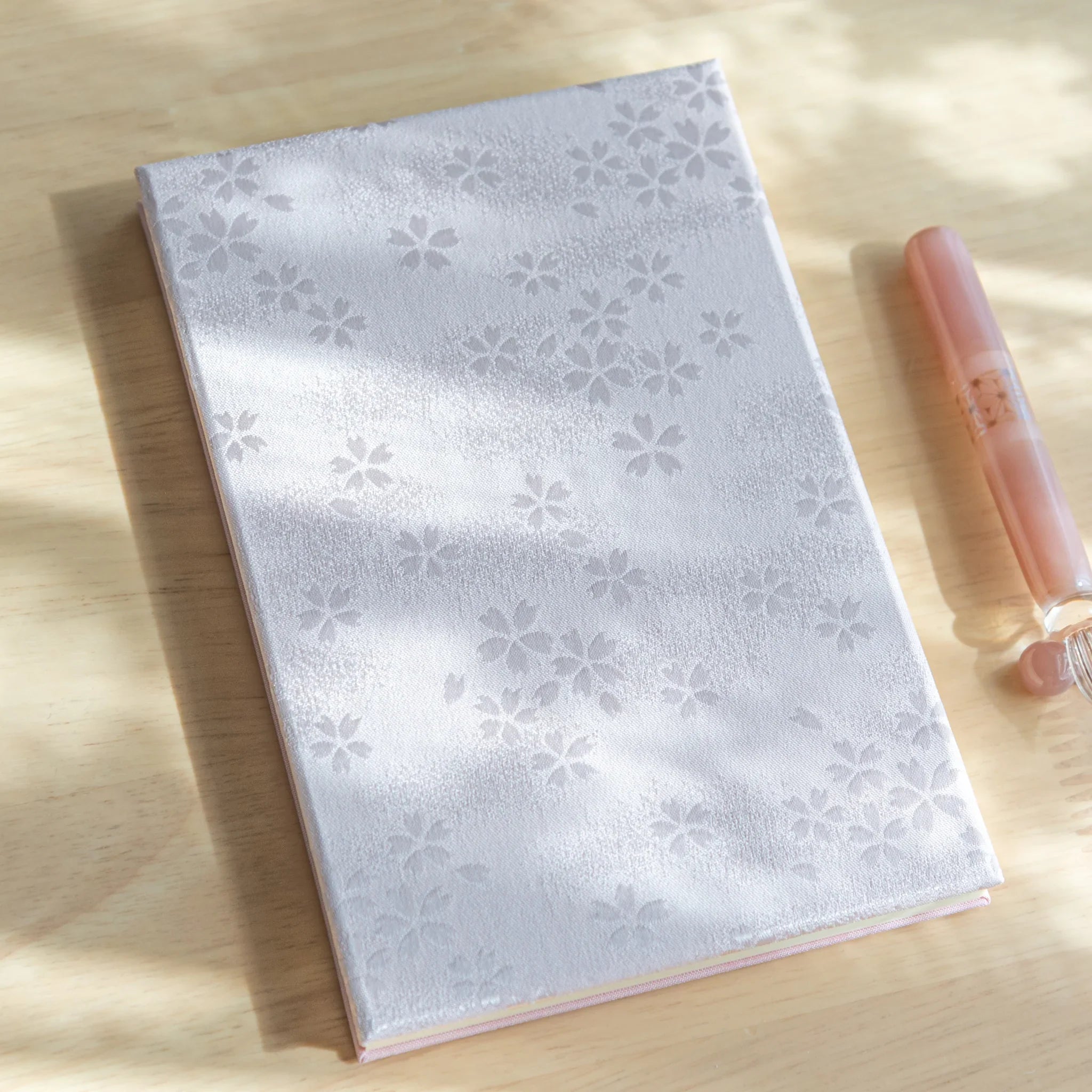 Sakura washi goshuincho front cover in silver with tone-on-tone sakura pattern on light wood, beside a peach glass dip pen