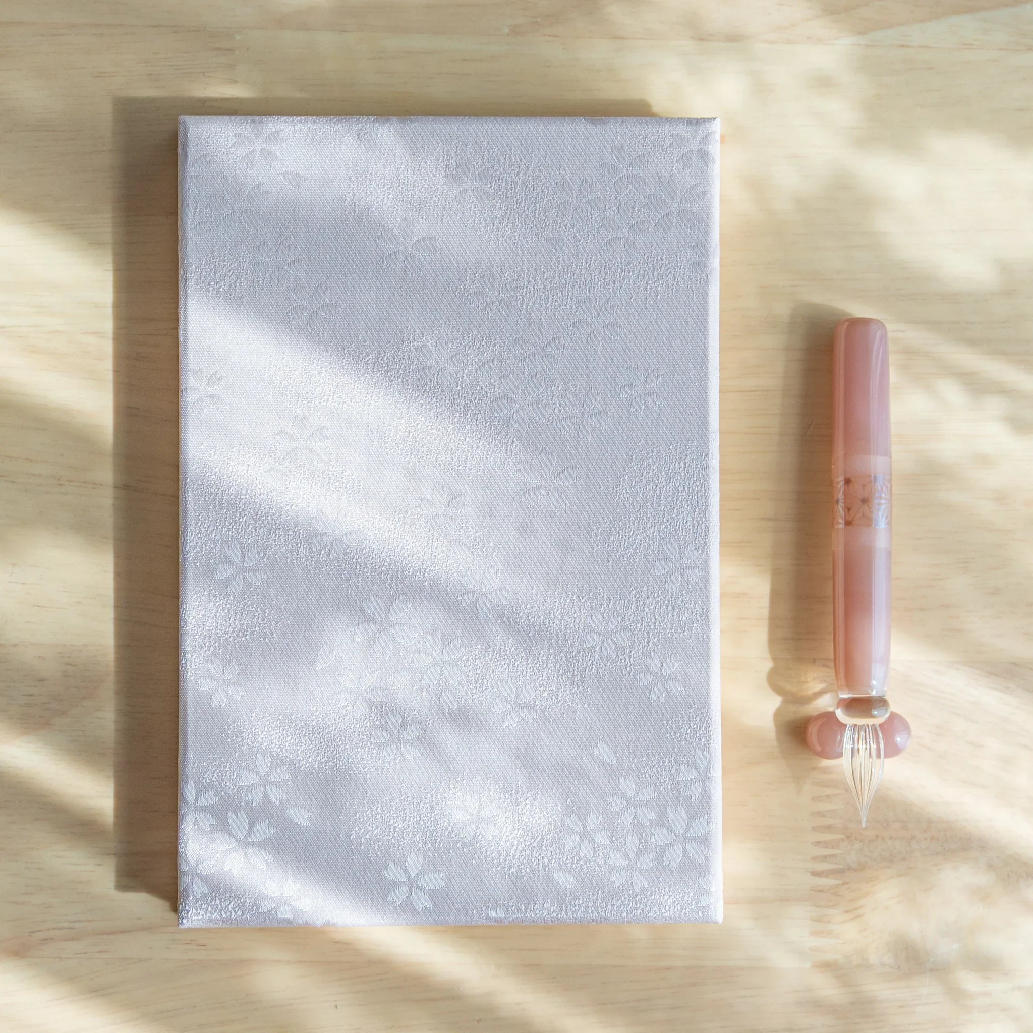 Sakura washi goshuincho front cover on light wood beside a peach glass dip pen in dappled sunlight