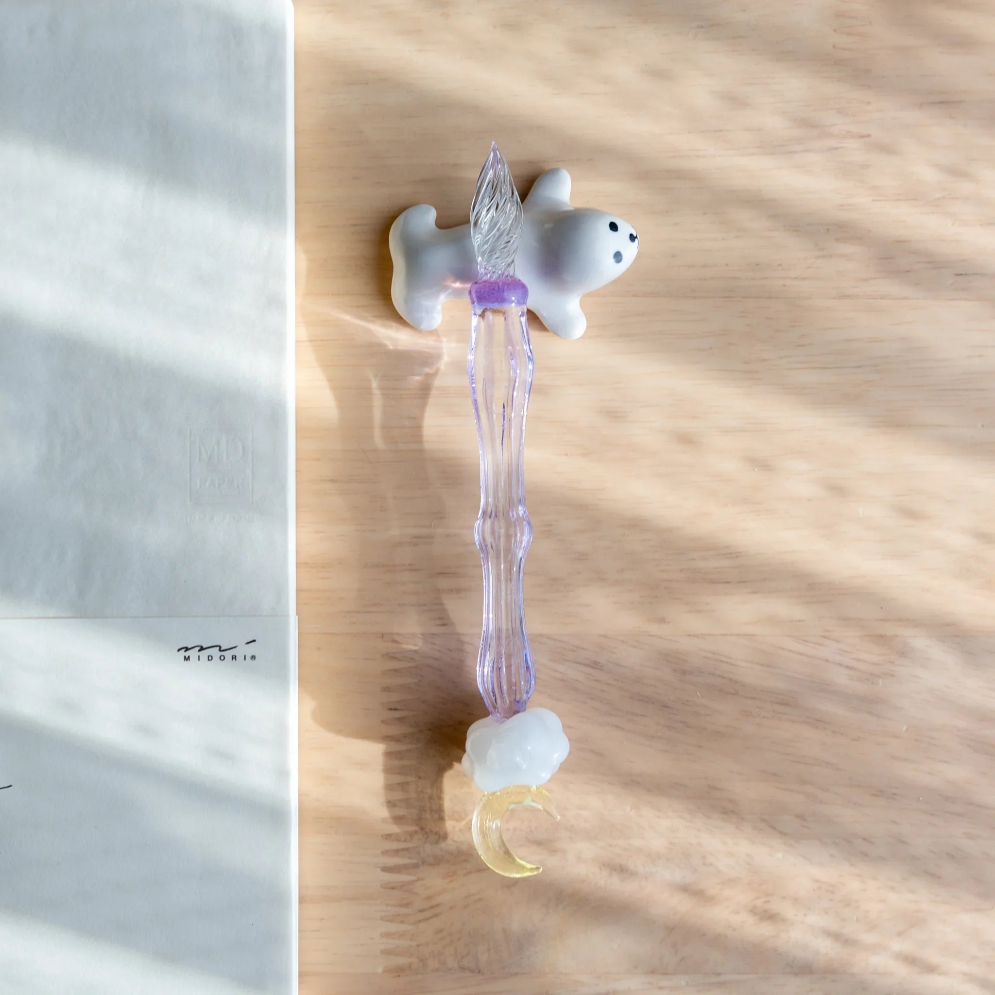Japanese glass dip pen with white ceramic seal figure, lavender barrel, cloud accent, and moon-shaped nib on wood