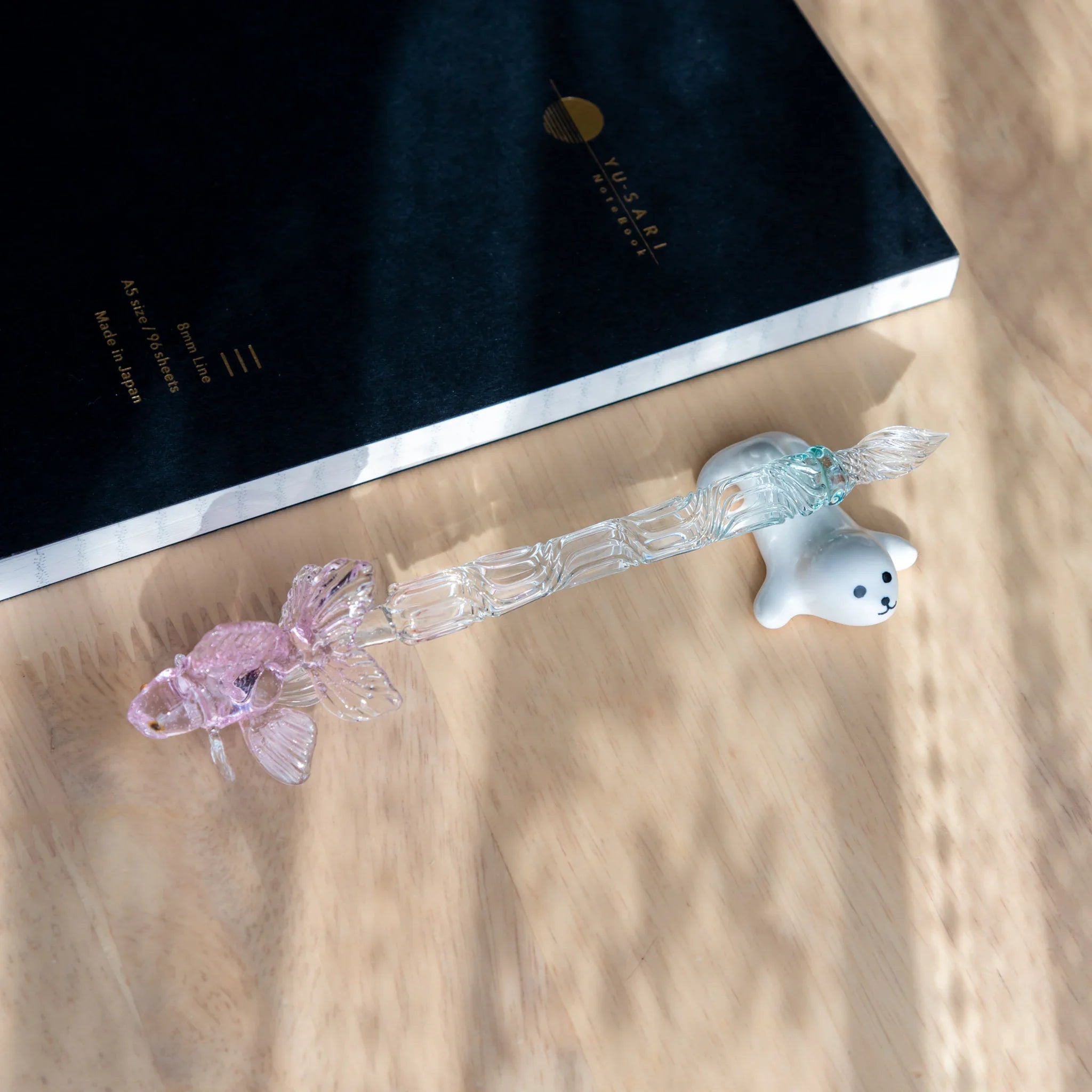 Japanese glass dip pen with ceramic seal handle, clear ribbed barrel, and pink glass fish nib, beside Yu-Sari notebook