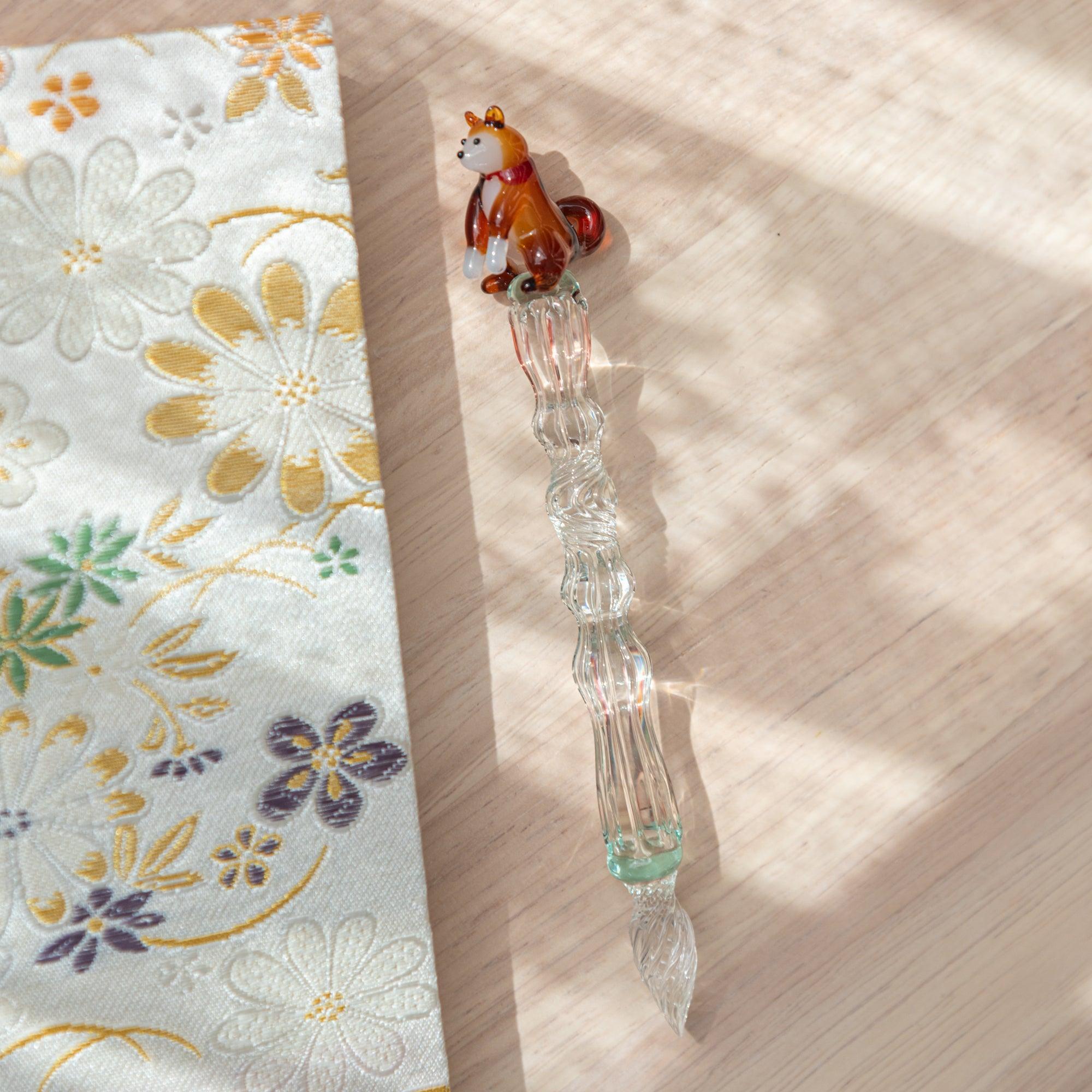Glass pen with Shiba Inu motif and clear light green shaft, displayed on a Kyoto Nishijin-ori notebook with floral patterns. Perfect for Japanese stationery lovers.