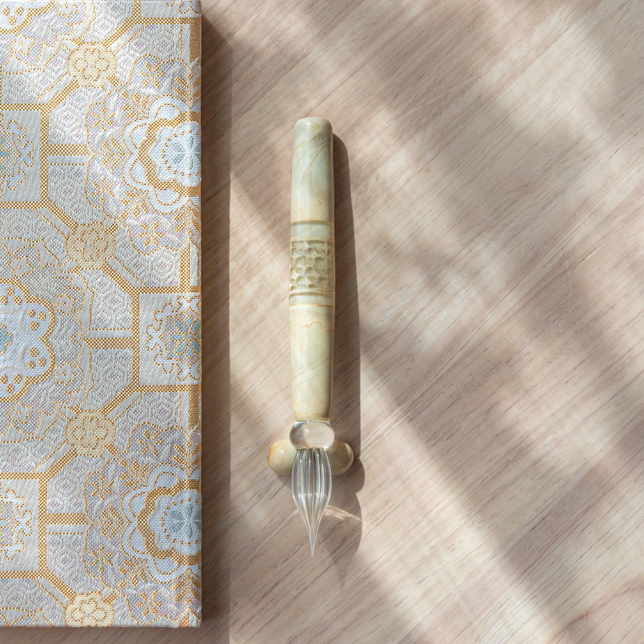 Ivory glass pen with a shippo design, paired with a matching glass pen rest. Displayed on a wooden surface beside a Nishijin textile notebook.