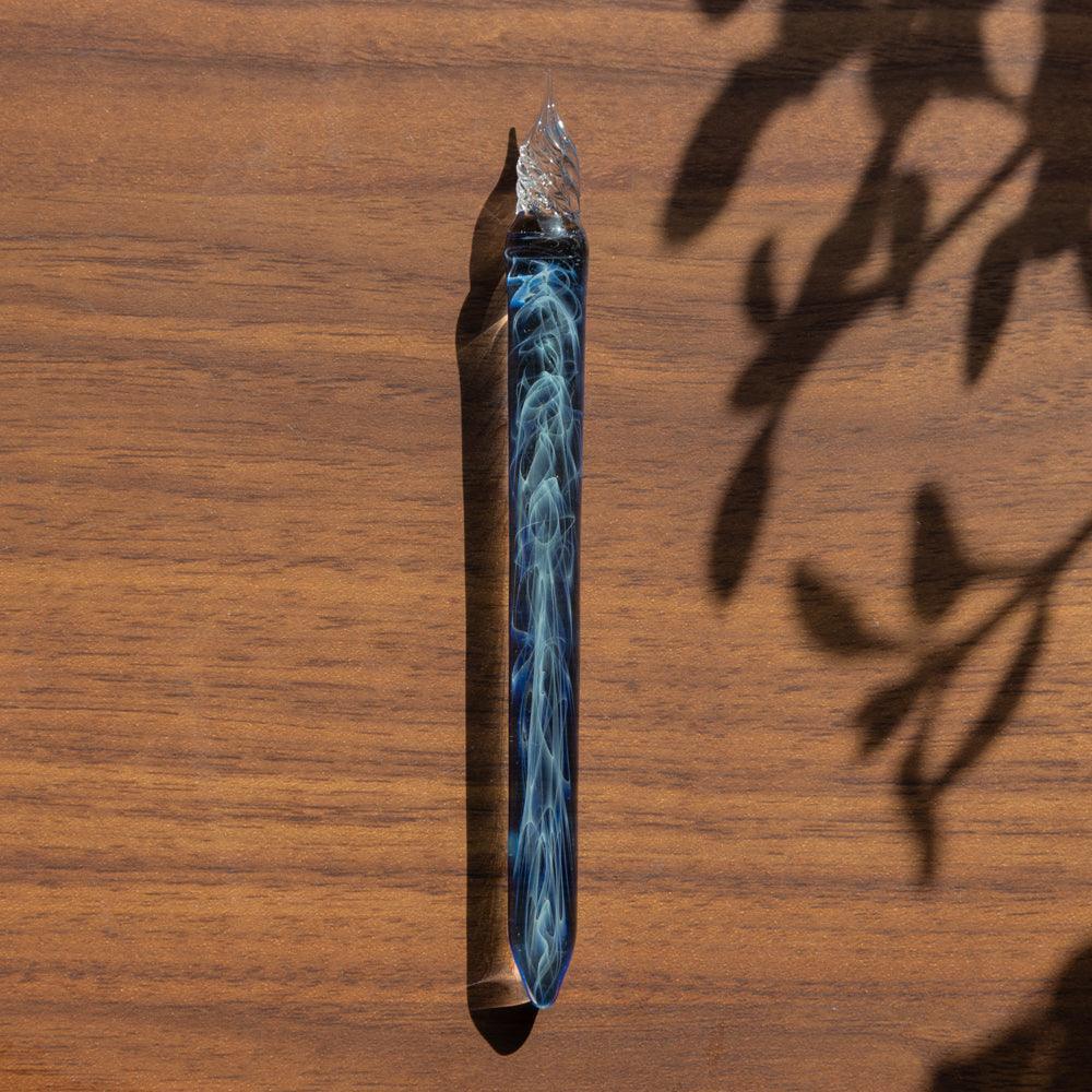 Blue smoky glass dip pen standing upright on wooden surface with leaf shadow cast on light background view