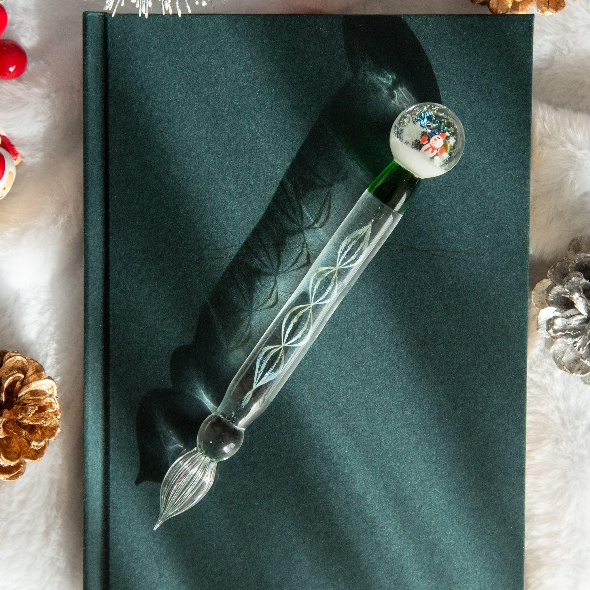 A Christmas-themed glass dip pen with a snow globe handle and intricate designs, resting on a dark green notebook with festive decorations.
