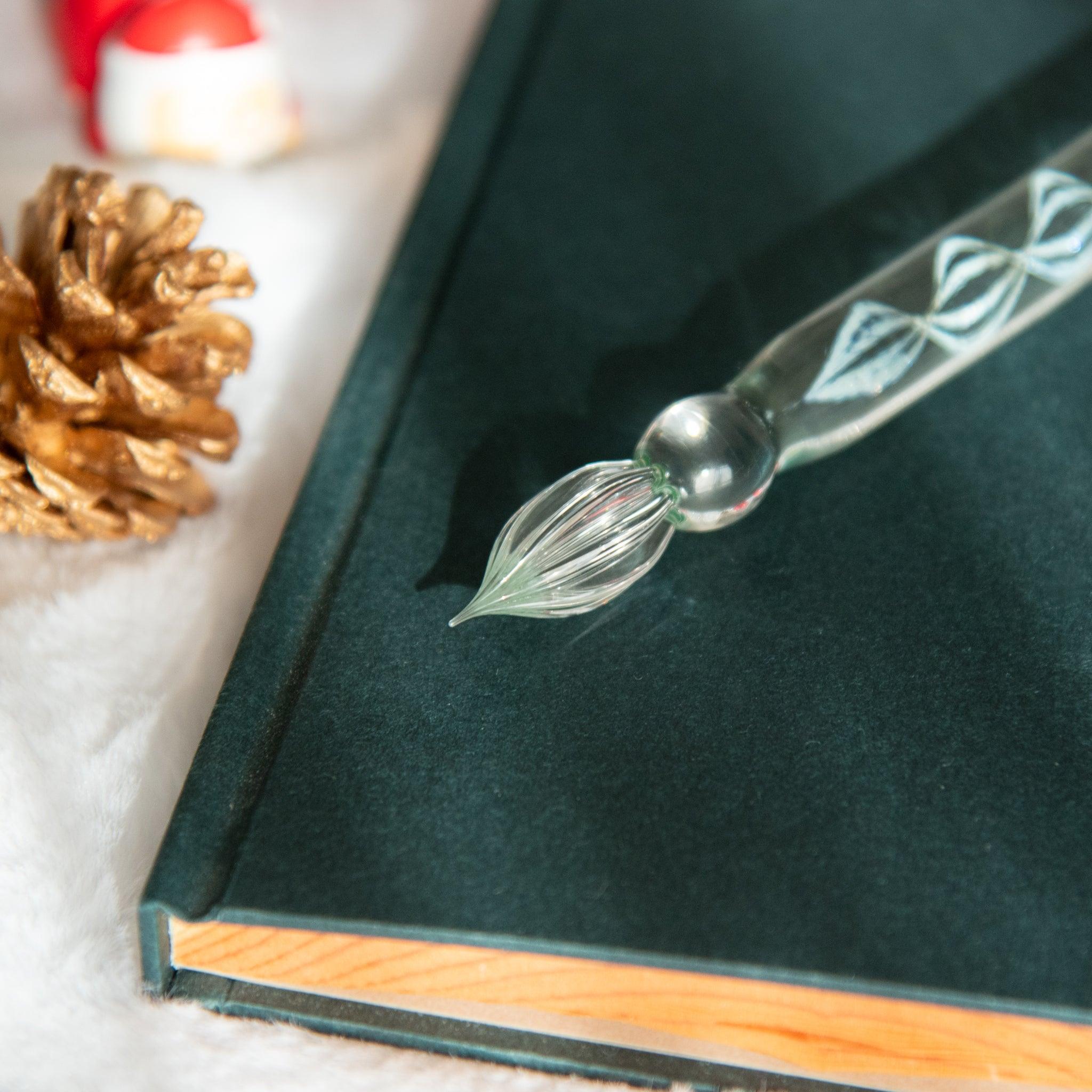 Close-up of a glass dip pen's nib with spiral engravings, placed on a dark green notebook surrounded by golden pinecones and festive accents.