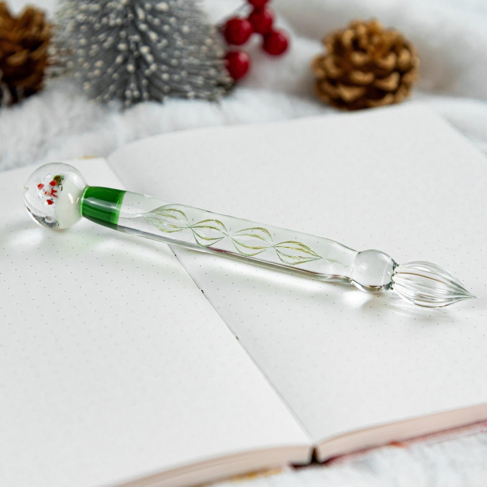 A Christmas glass dip pen featuring a snow globe handle with a Santa design, lying on an open dotted notebook with festive background elements.