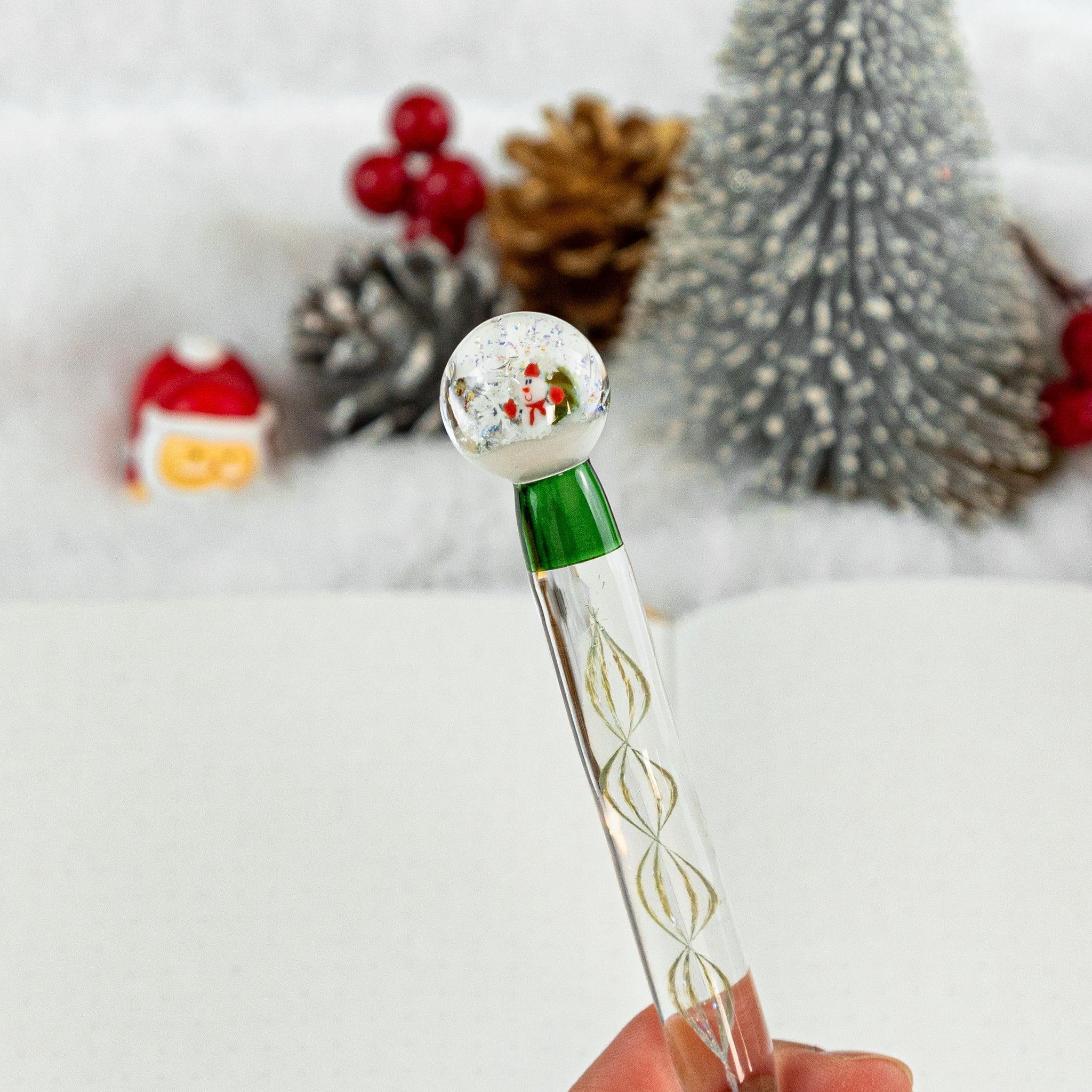 The handle of a glass dip pen shaped as a snow globe with Santa inside, held above an open dotted notebook with a winter-themed background.