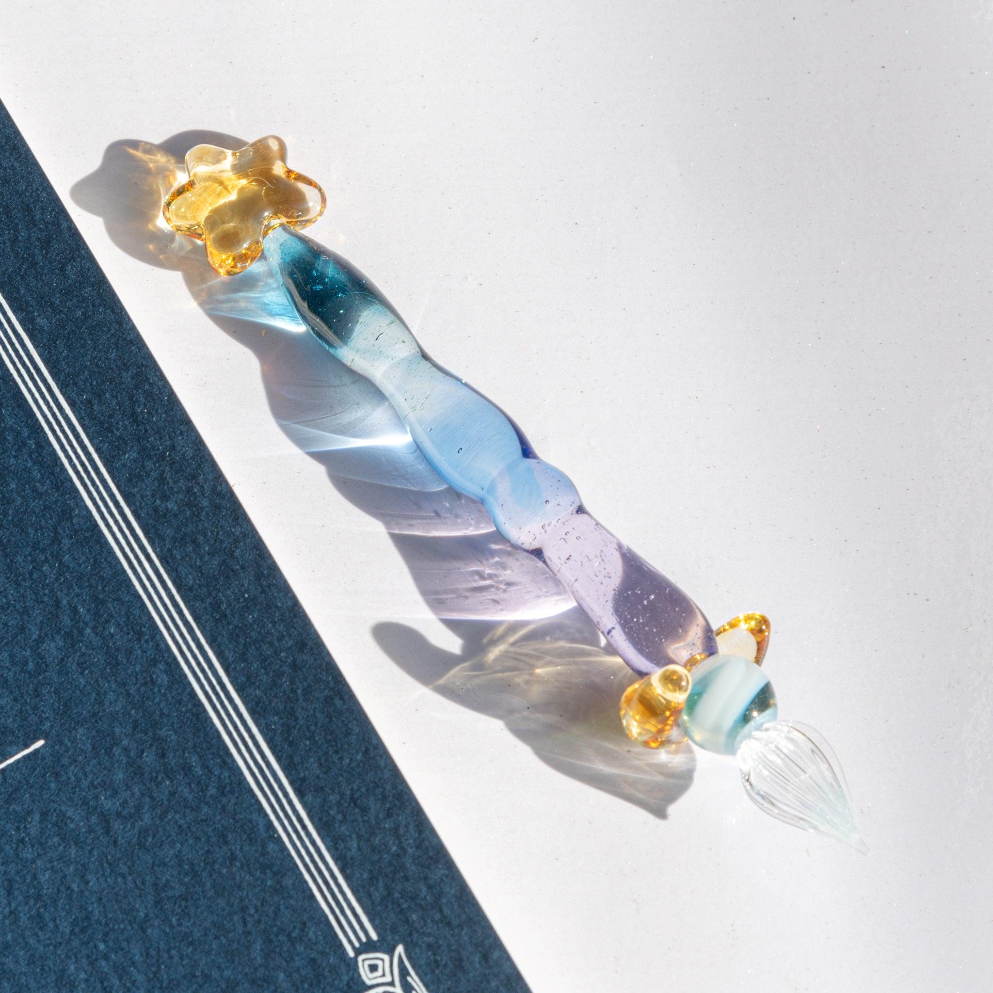 Glass dip pen with blue-purple gradient, gold star top, and clear nib, casting light reflections. Japanese stationery.