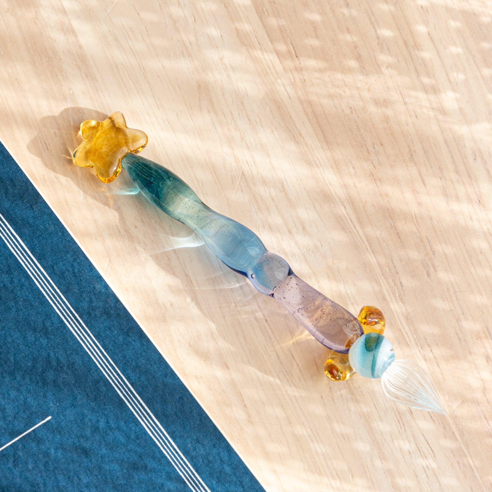Glass dip pen with a blue-purple gradient body, gold star top, and clear nib, placed on a wooden surface.