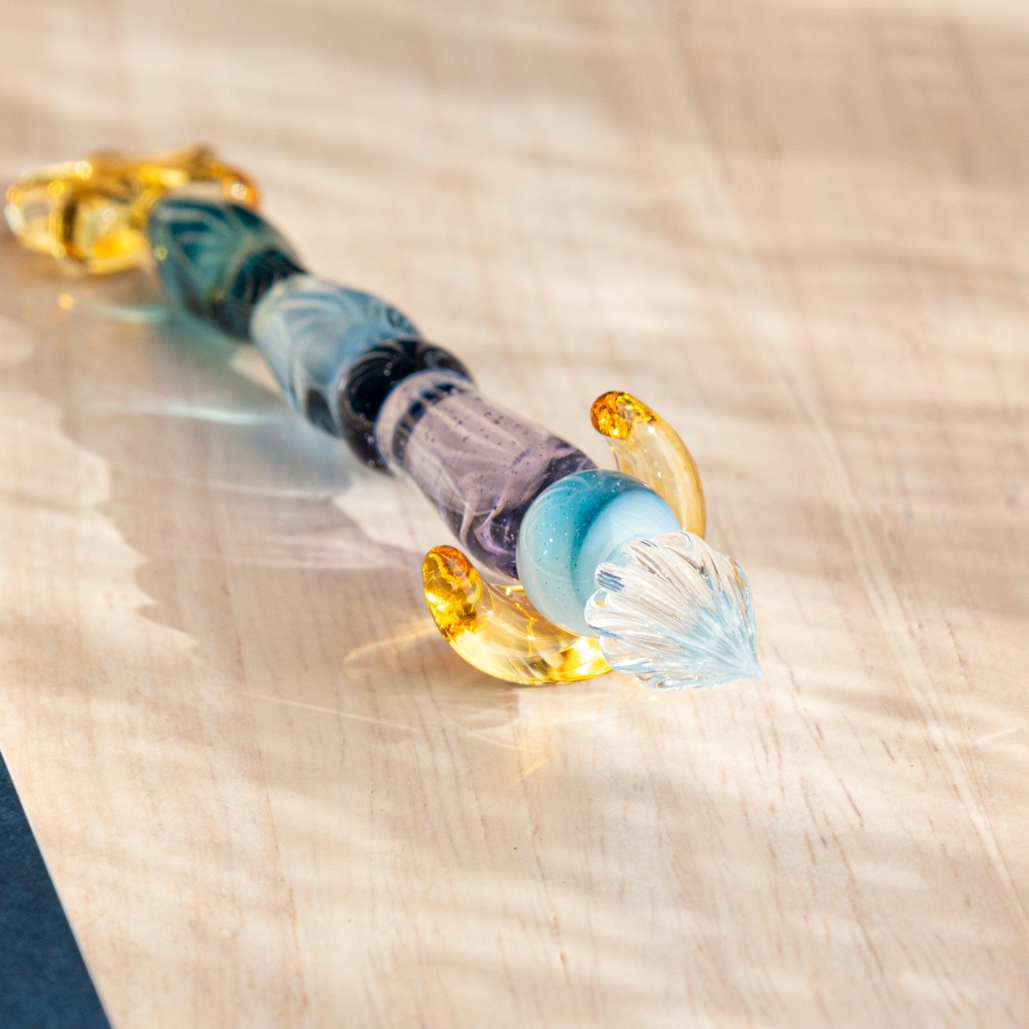 Close-up of a glass dip pen with a twisted blue-purple body, clear nib, and two gold crescent accessories on wood.