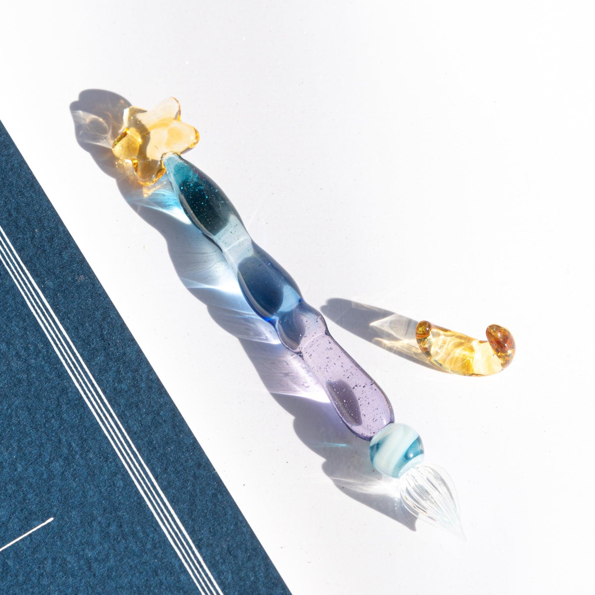Glass dip pen with a twisted blue-purple body, clear nib, and a detached gold crescent accessory on white paper.