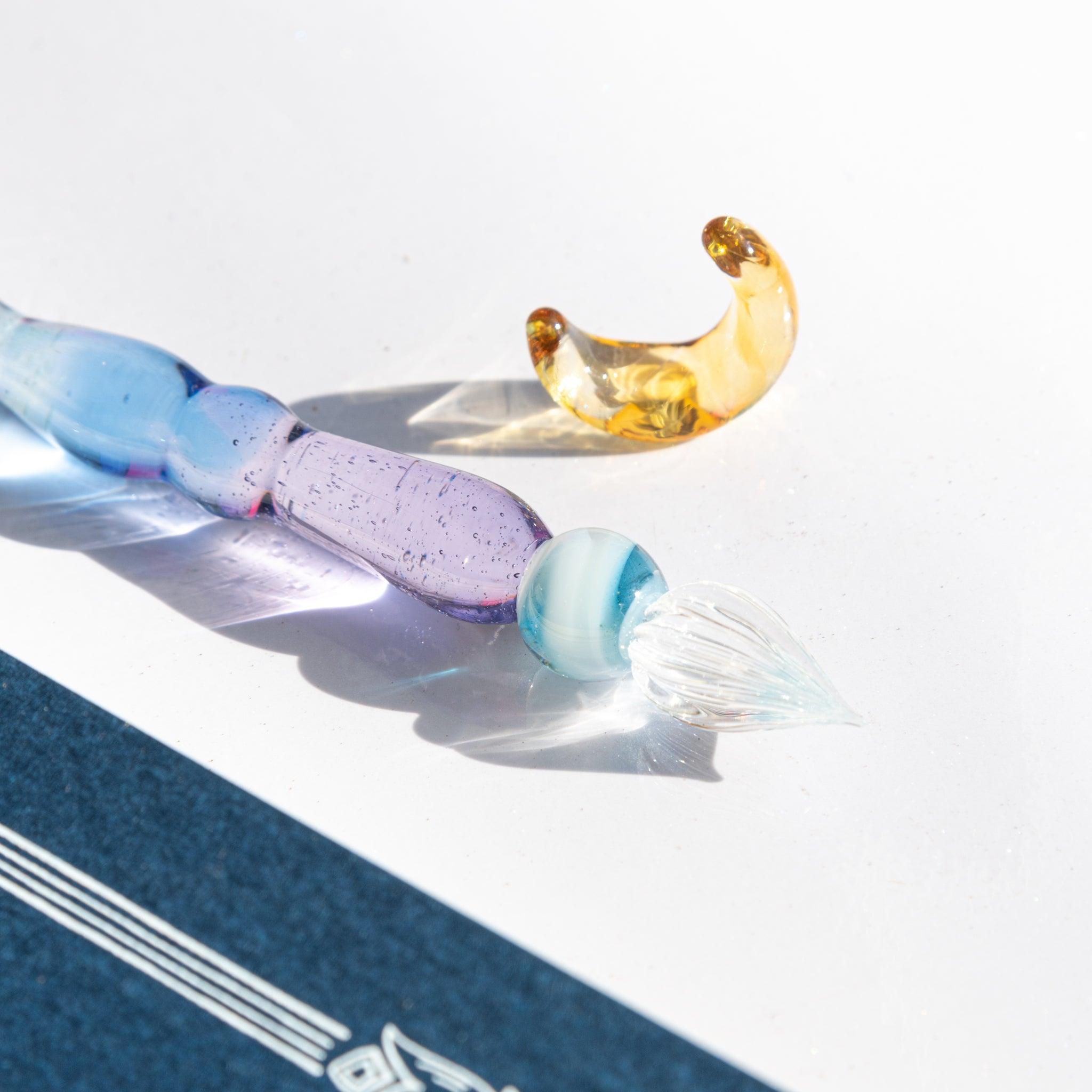 Close-up of a glass dip pen with a clear nib and a gold crescent accessory, reflecting light.