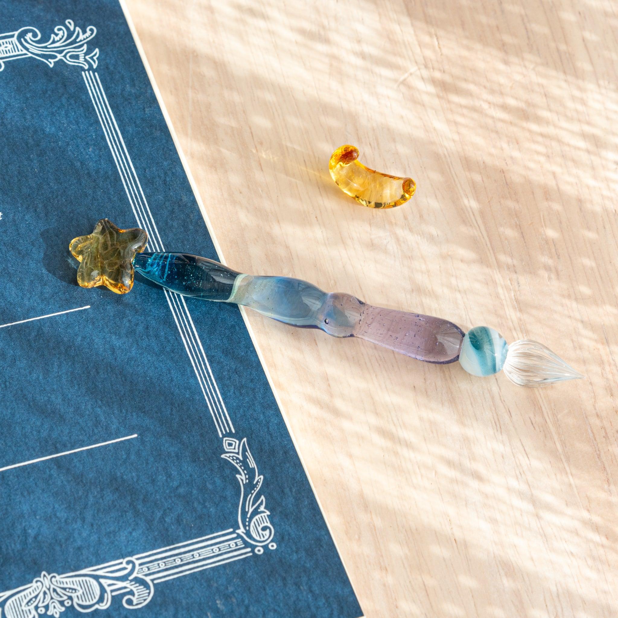 Glass dip pen with a gold star top and detached crescent accessory, resting on a wooden surface with a blue notebook.