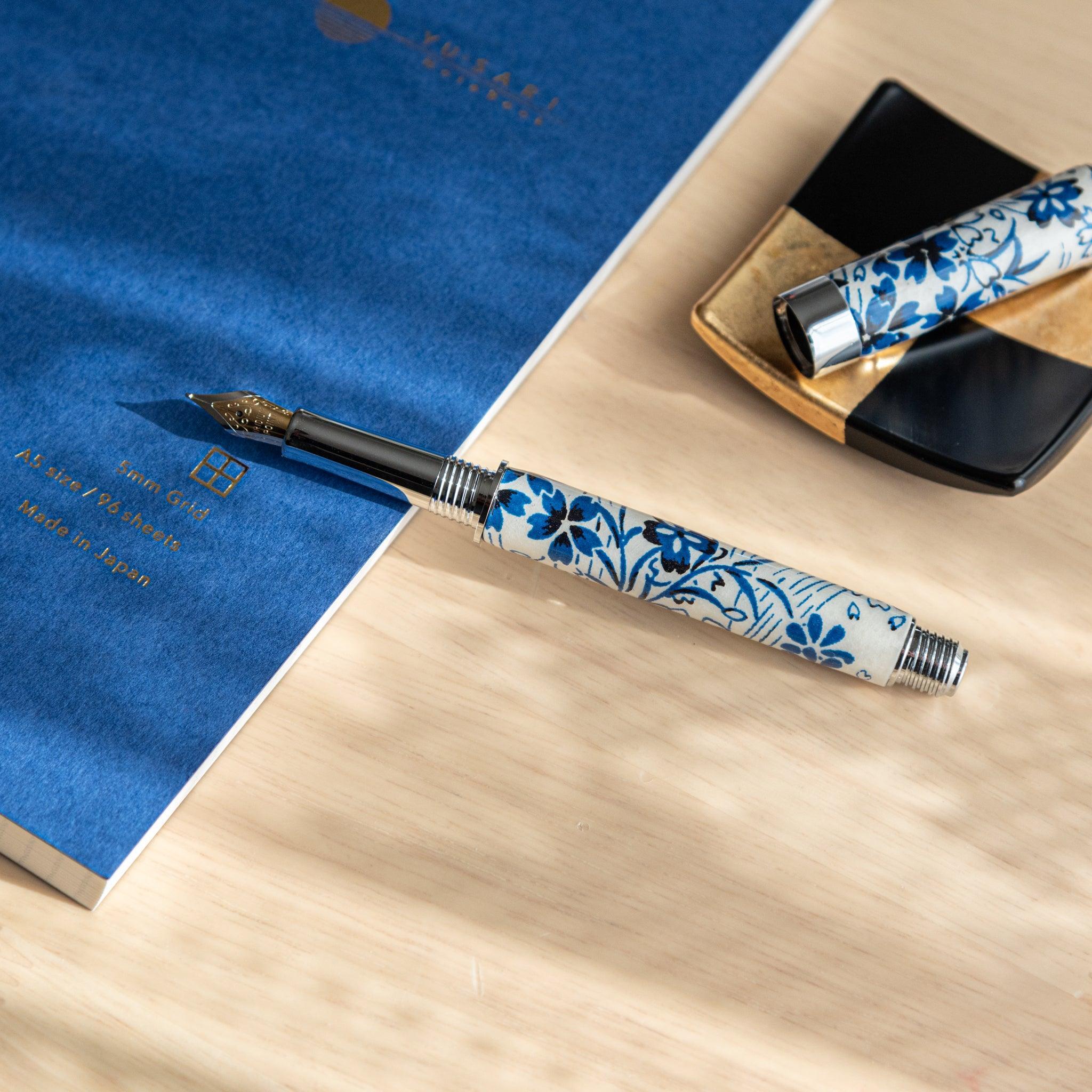 A Japanese stationery fountain pen with blue floral designs, uncapped, placed diagonally on a wooden surface near a blue notebook.