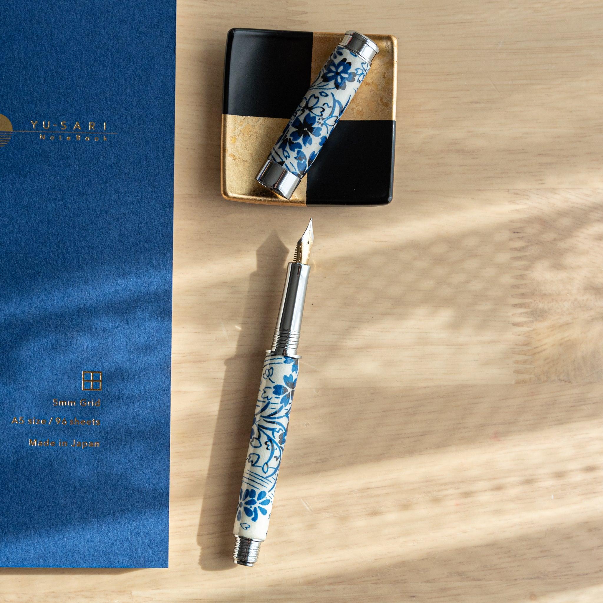 A Japanese stationery fountain pen with blue floral patterns, disassembled, displayed alongside a blue notebook and decorative dish.