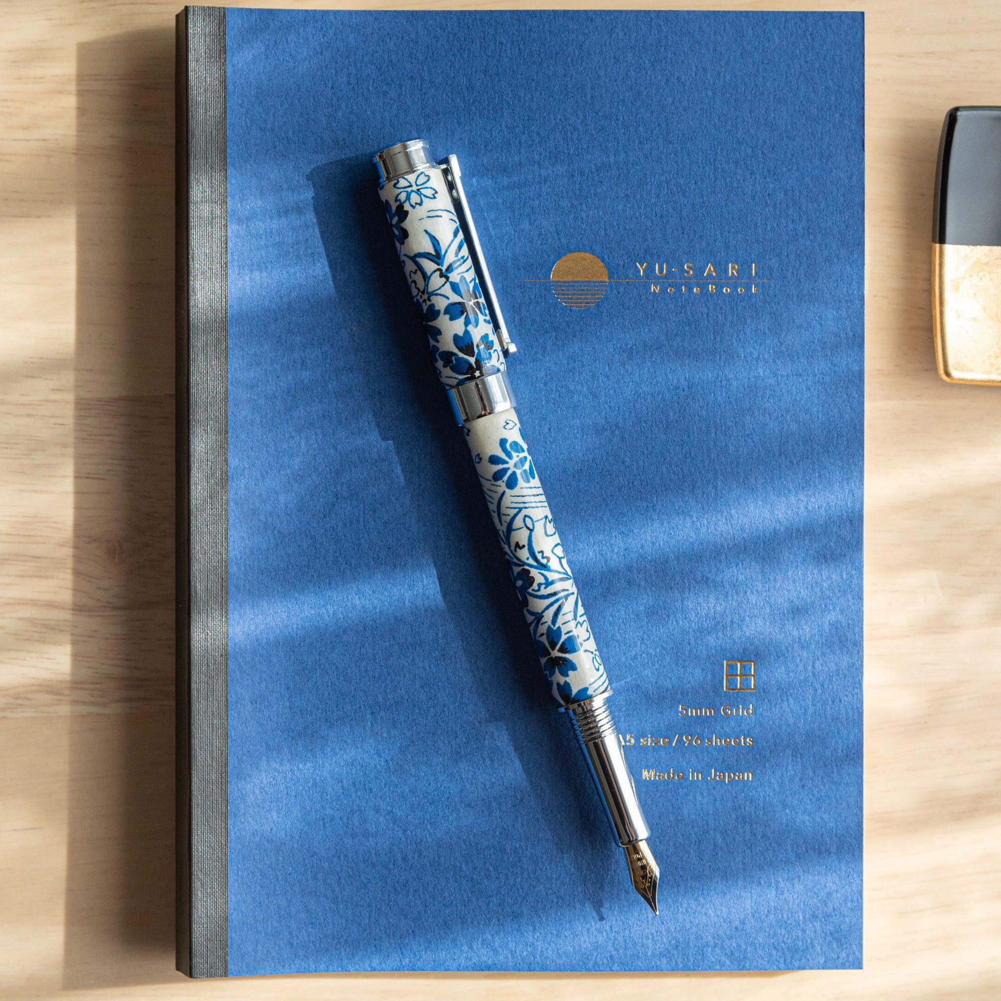 A Japanese stationery fountain pen with blue floral details and a silver nib, resting on a blue notebook under soft light.