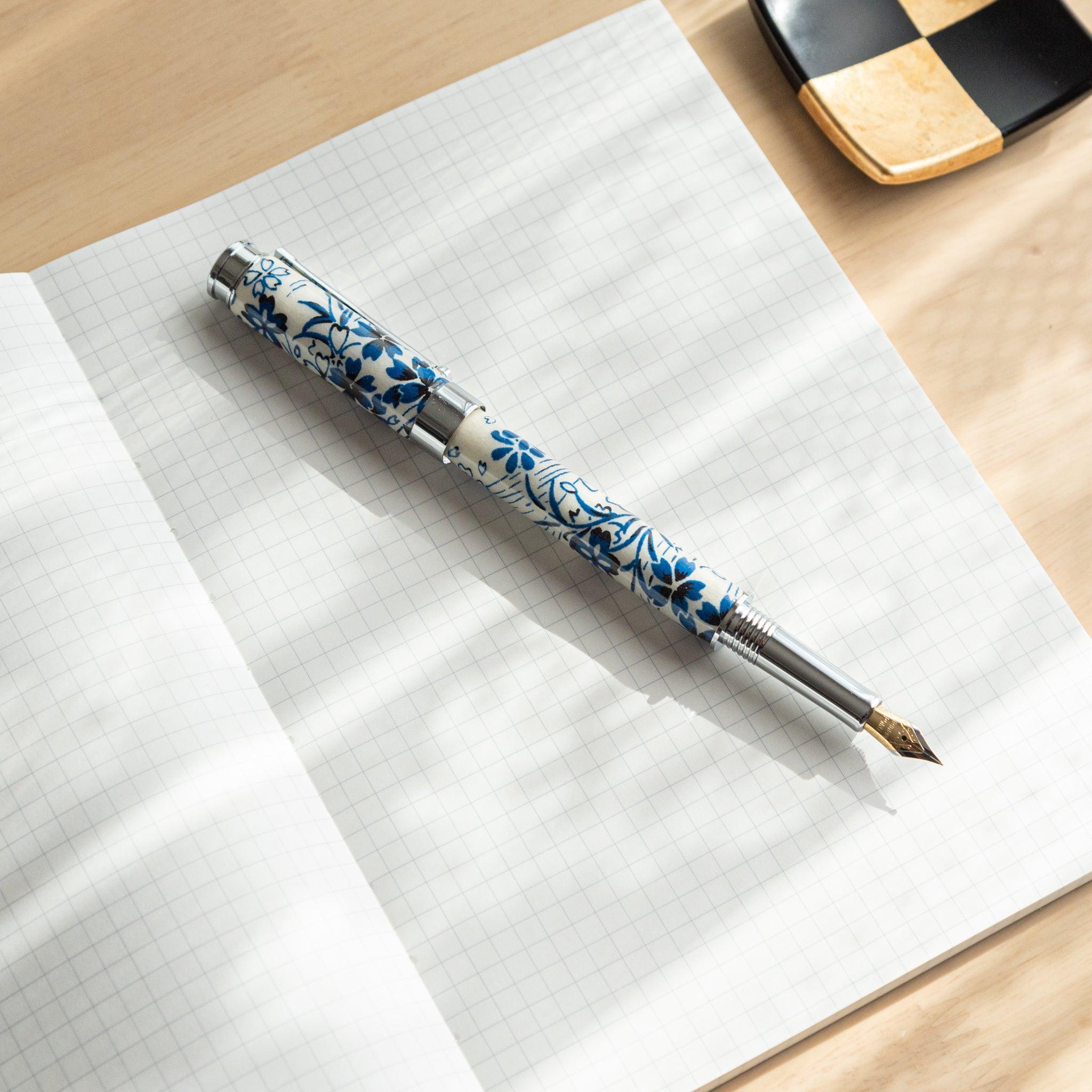 A Japanese stationery fountain pen with blue floral patterns and a silver nib, placed on an open grid notebook under soft light.