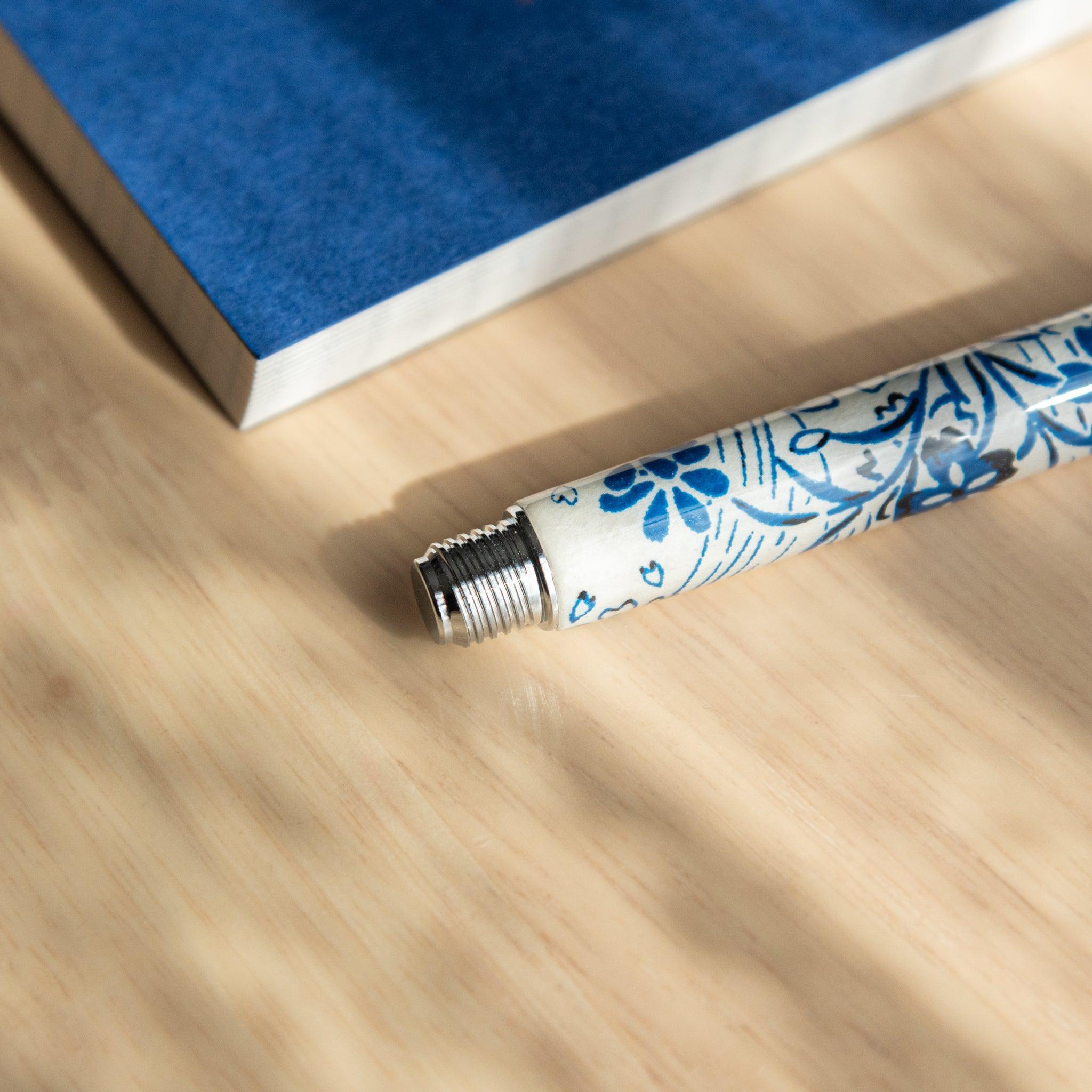 A Japanese stationery fountain pen barrel with blue floral details, showing its threaded end, placed near a blue notebook on a wooden surface.