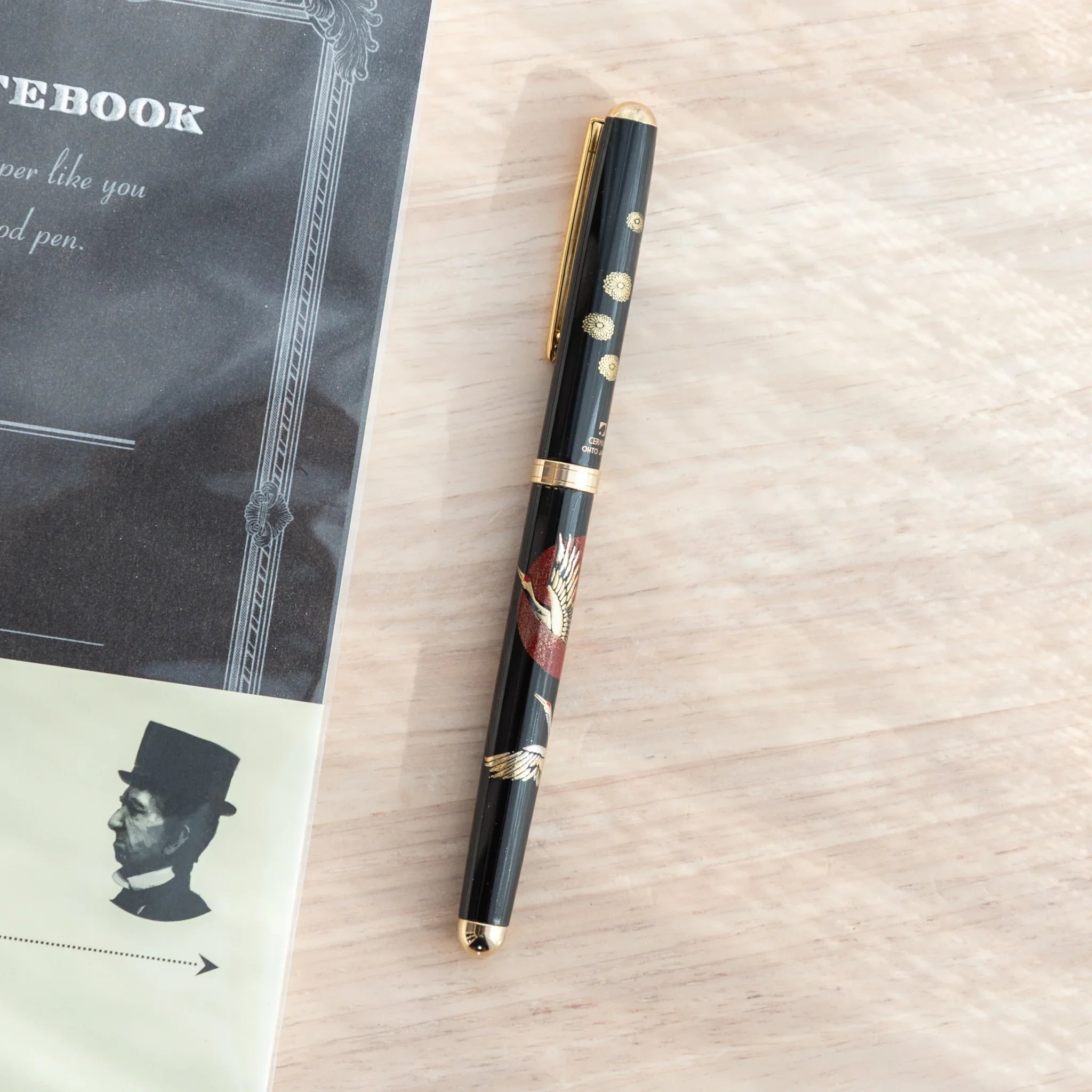 Black lacquer Japanese ballpoint pen with white crane and red sunrise pattern, gold chrysanthemum dots, on light wood