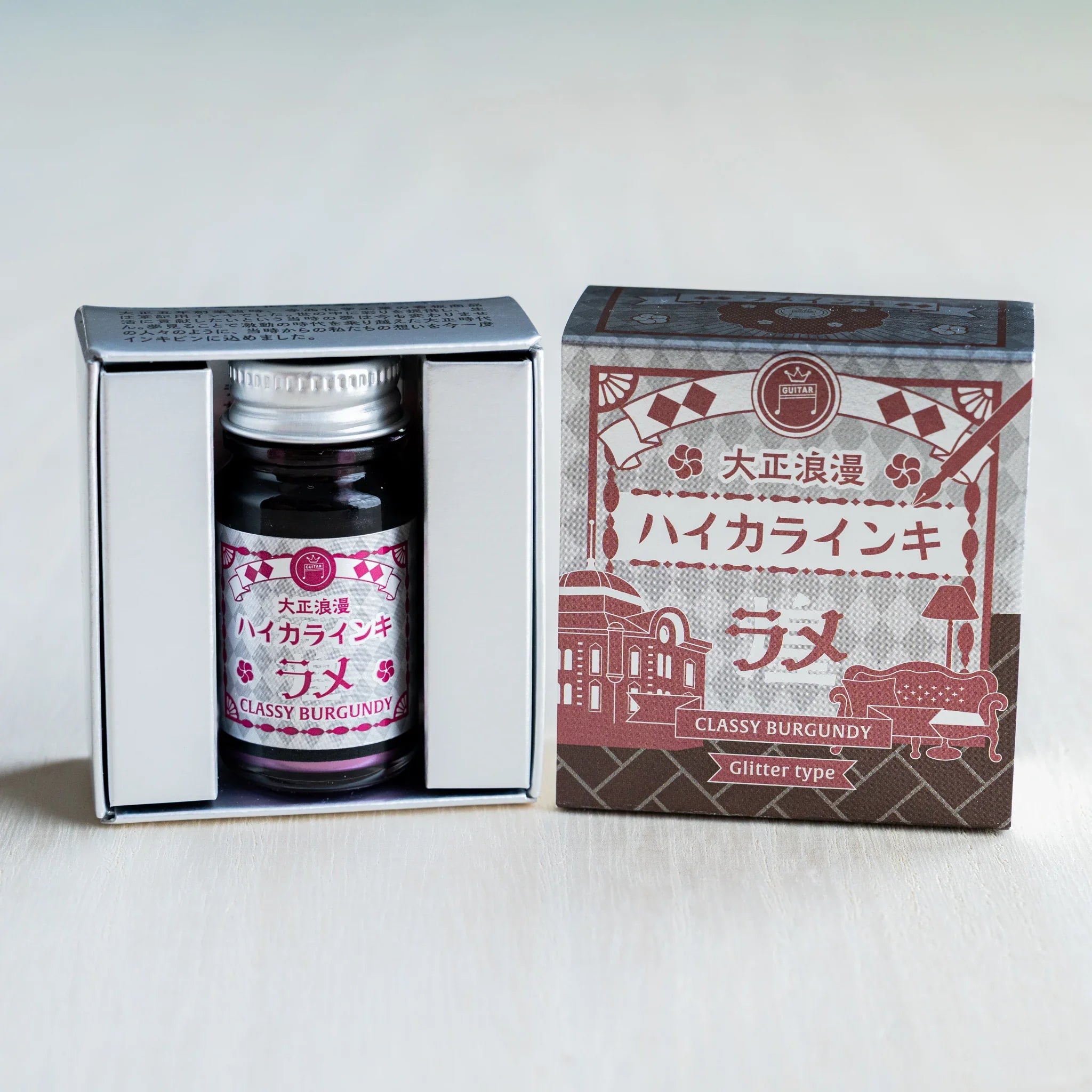 Taisho Romance Classy Burgundy Shimmer Ink bottle in its open black inner box beside the closed silver foil outer box