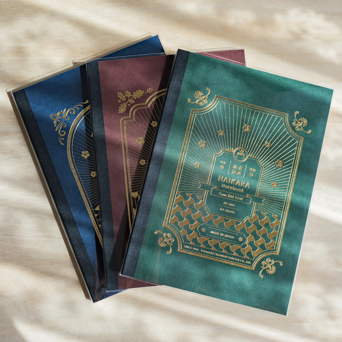 Three HAIKARA notebooks in dark blue, dark red and dark green, each with a gold foil frame design on the cover, overlapping