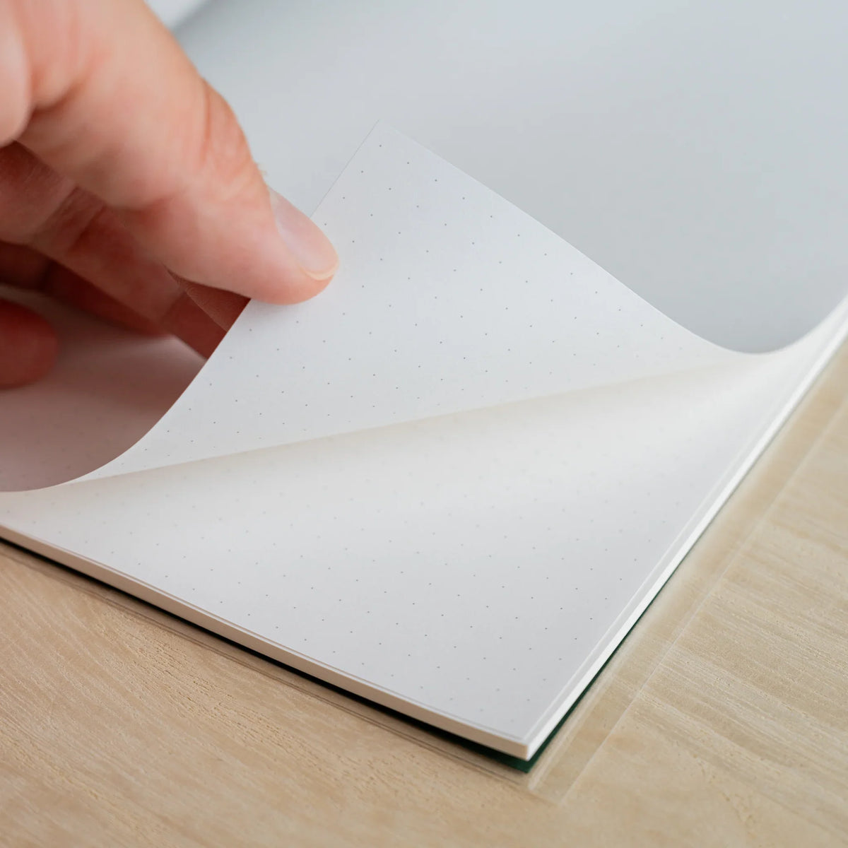 Hand turning a dot grid page of the HAIKARA notebook, with the dark green cover visible under the stacked sheets