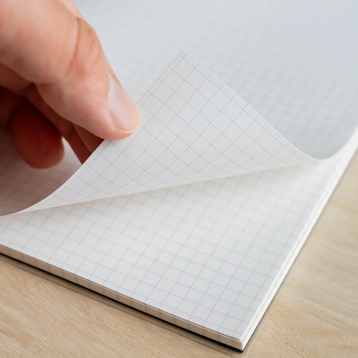 Hand turning a 5mm grid page of the HAIKARA notebook, showing the grey ruling and the stacked block of sheets