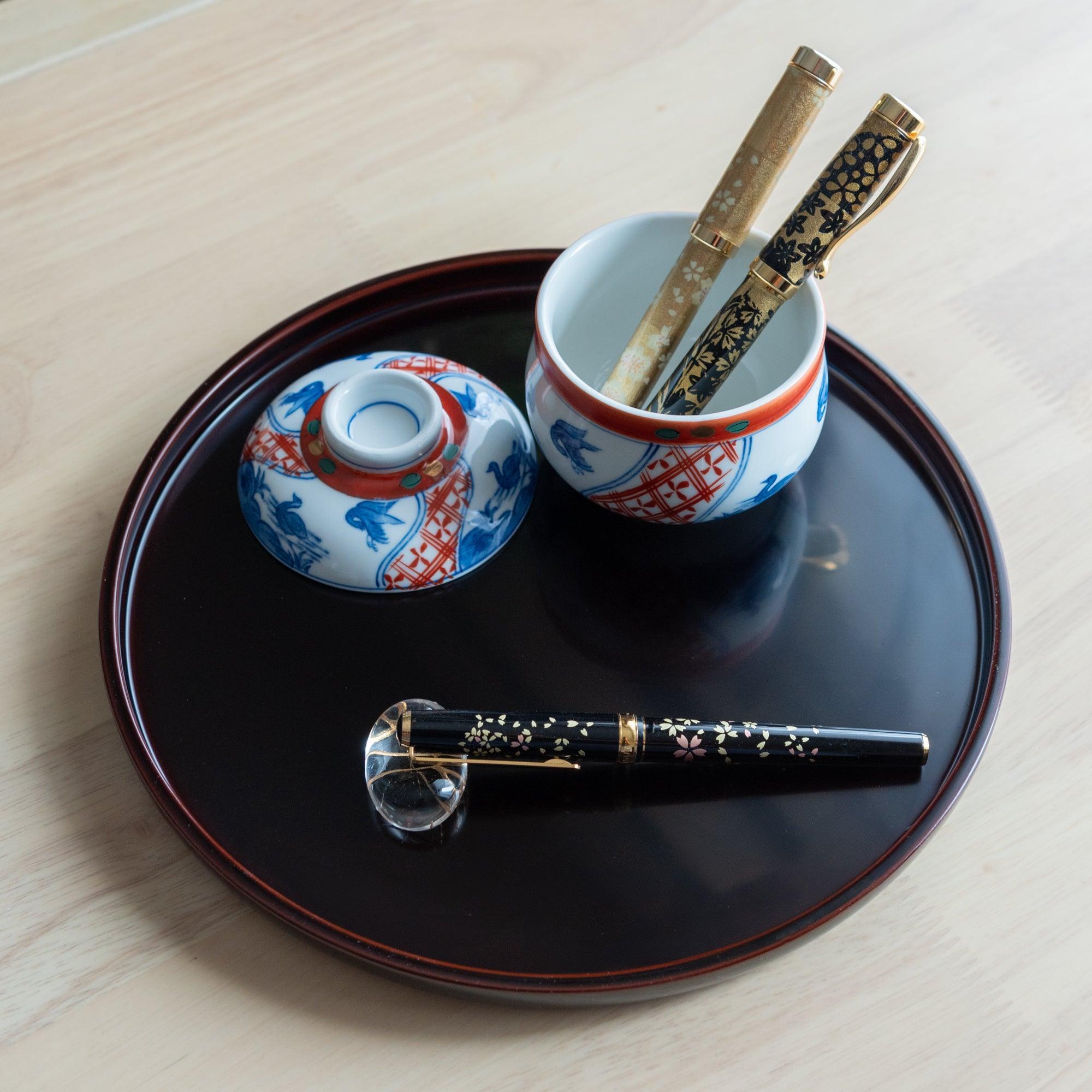 Two gold fountain pens inside a Japanese porcelain jar with a sakura-themed pen placed below, arranged on a black lacquered tray.