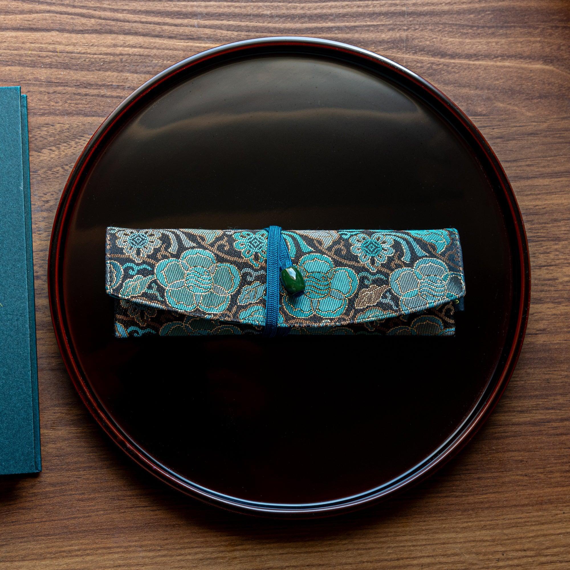 A compact Japanese stationery pen case with a turquoise floral brocade pattern and green bead clasp, displayed on a black lacquered tray.