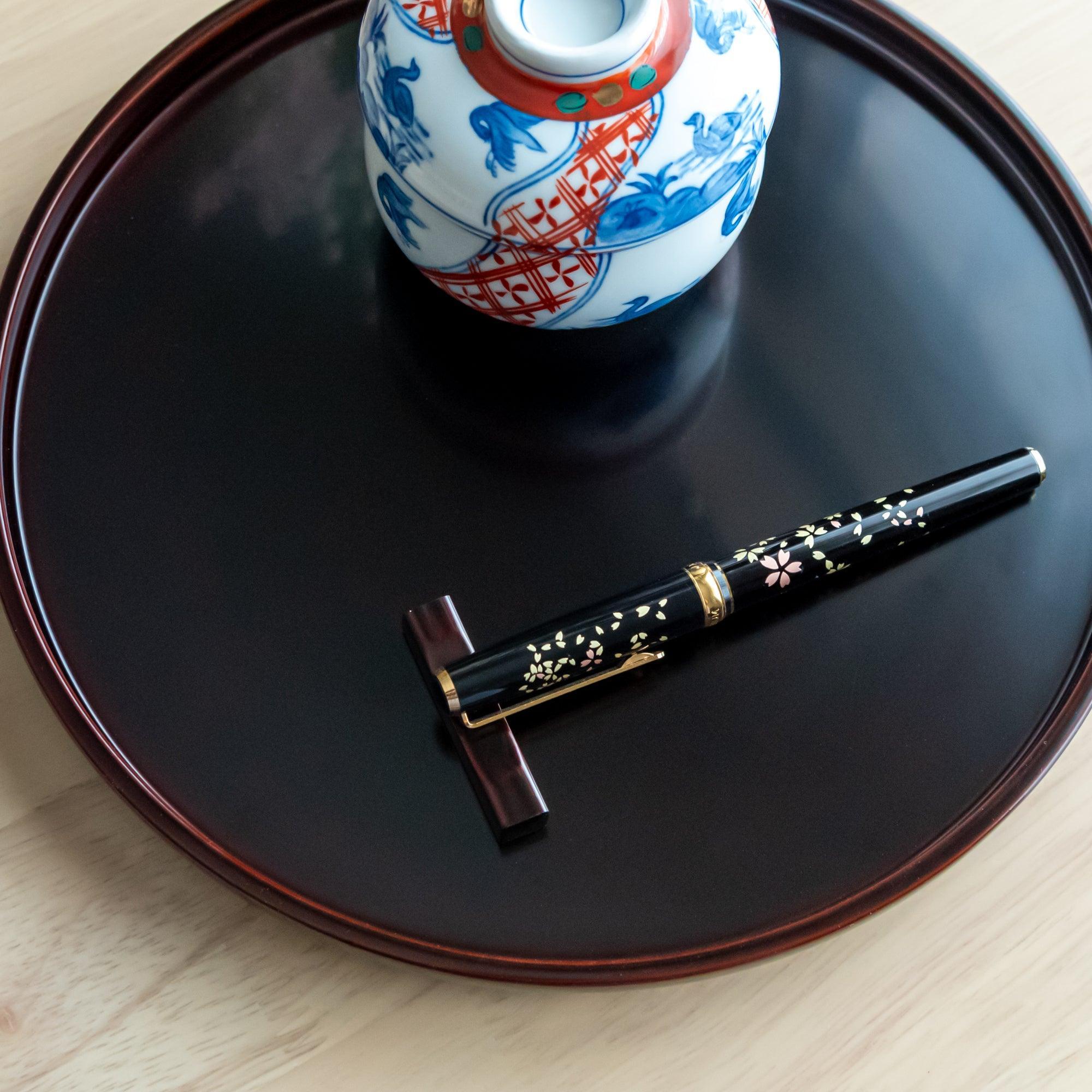 A black fountain pen with sakura motifs, part of Japanese stationery, lies on a red pen rest on a glossy black lacquer tray.