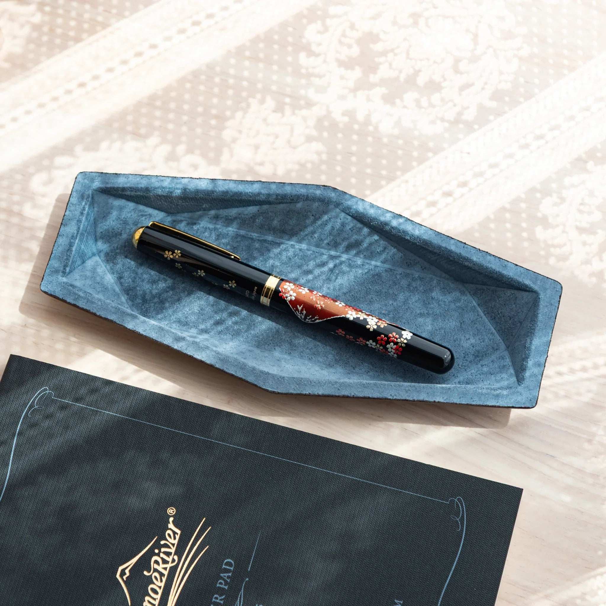 Blue-gray suede hexagonal slim pen tray holding Japanese fountain pen with red floral pattern and gold trim, on white fabric