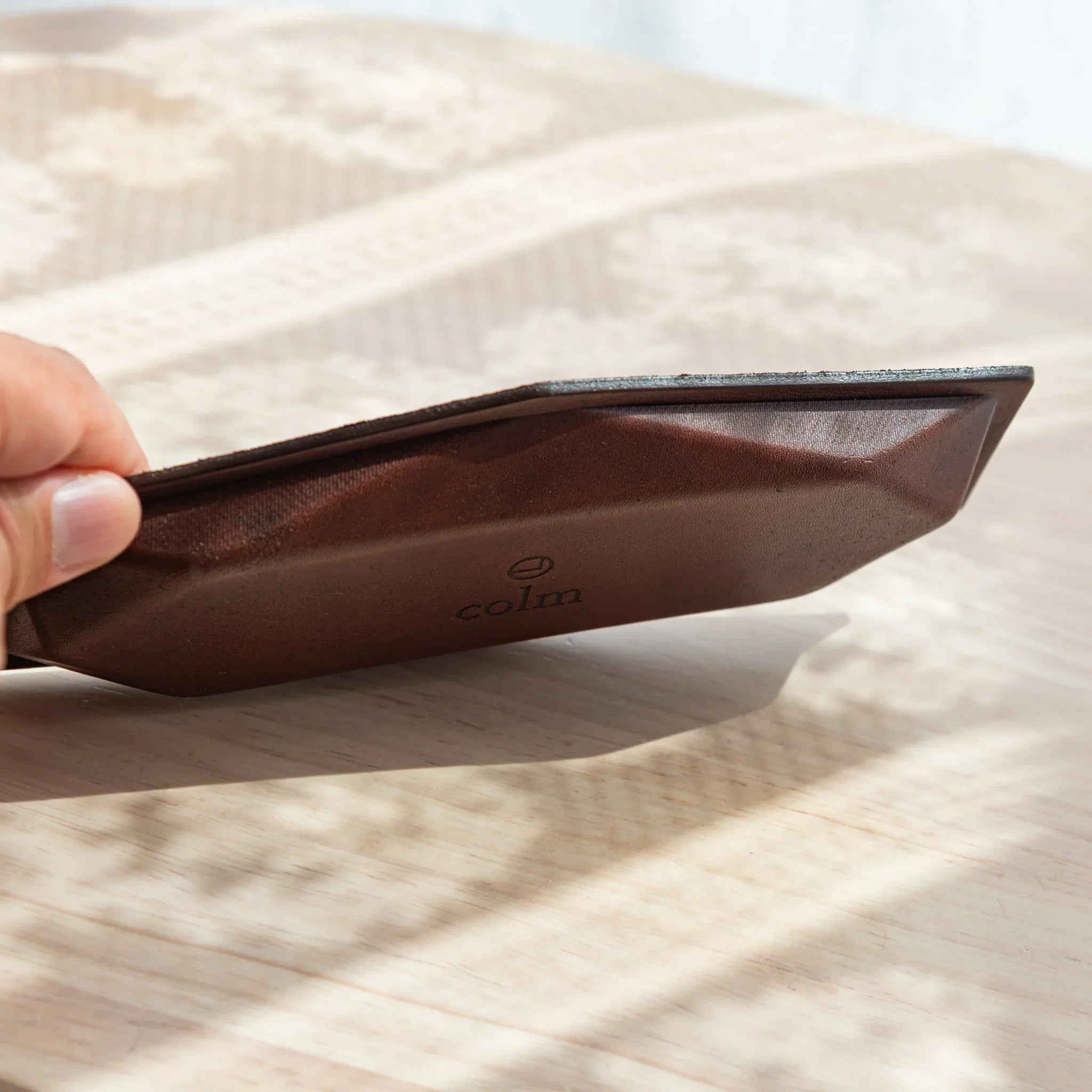 Hand tilting a hexagonal slim pen tray to show the brown leather base with embossed colm logo, blue-gray suede edge visible