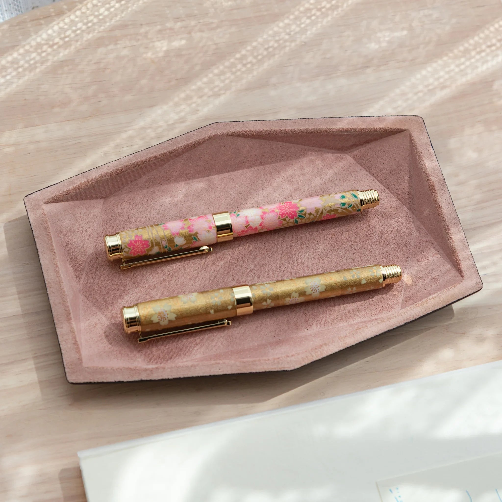 Pink suede wide hexagonal pen tray holding two Japanese floral fountain pens with gold trim, overhead view on white surface