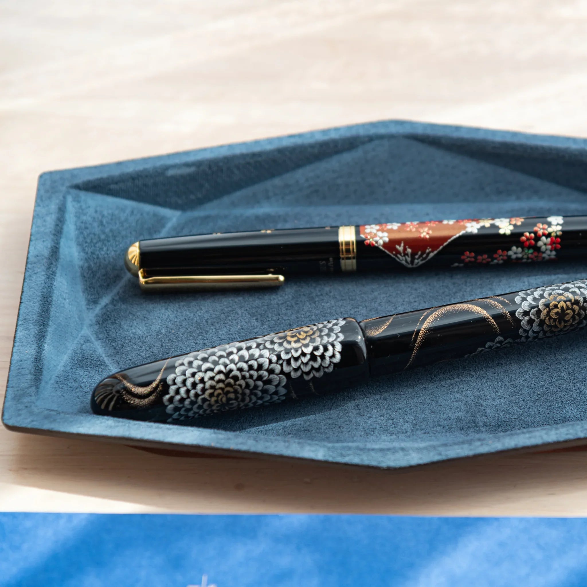 Two lacquered fountain pens placed on a blue Japanese Pen Tray made of suede and leather.