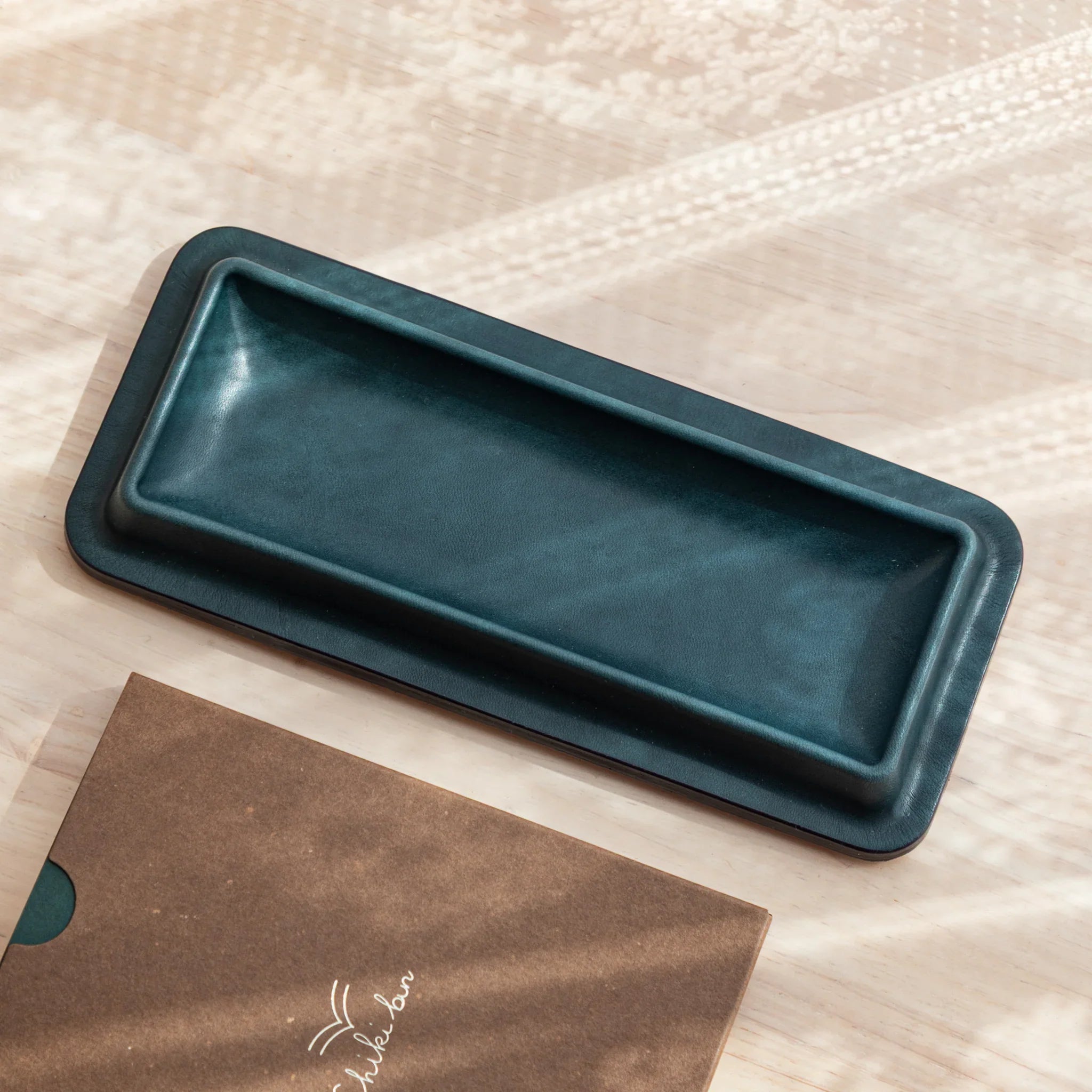 Tatsuno Leather Pen Tray Slim in dark green, shown empty beside a brown Shikibon notebook.